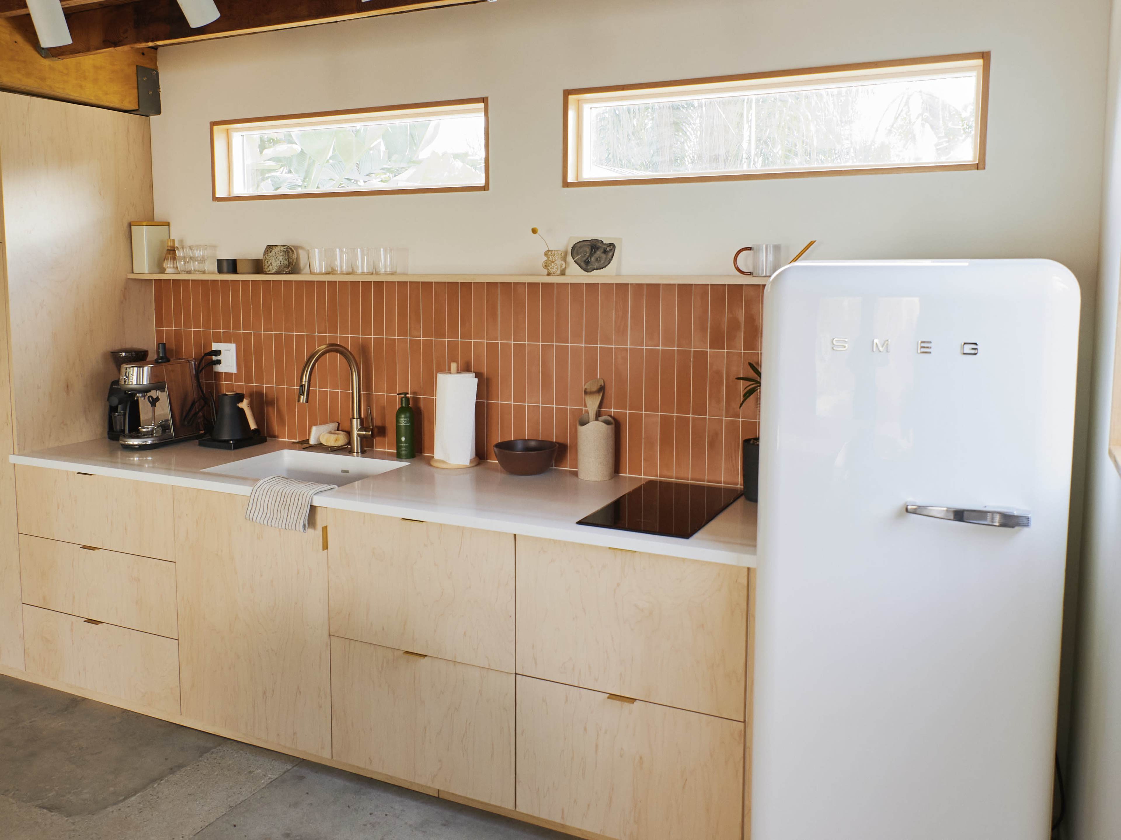 A modern kitchen features a light wooden cabinetry, a white SMEG refrigerator, and a pink tile backsplash with various kitchen essentials on the counter.