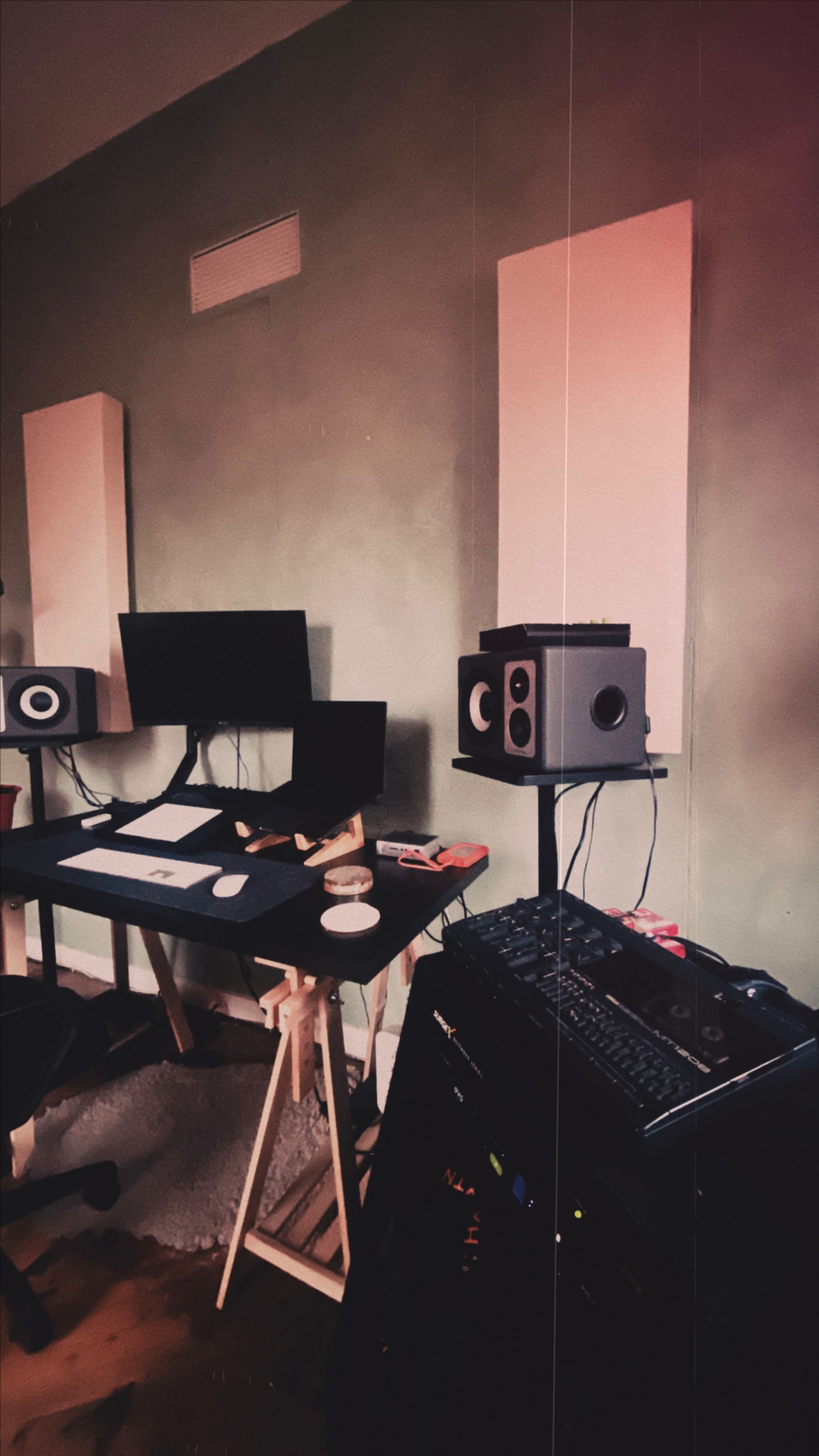 The image shows a corner of a music studio featuring a black desk with dual monitors, studio speakers mounted on the wall, and an audio mixer beside a computer setup.