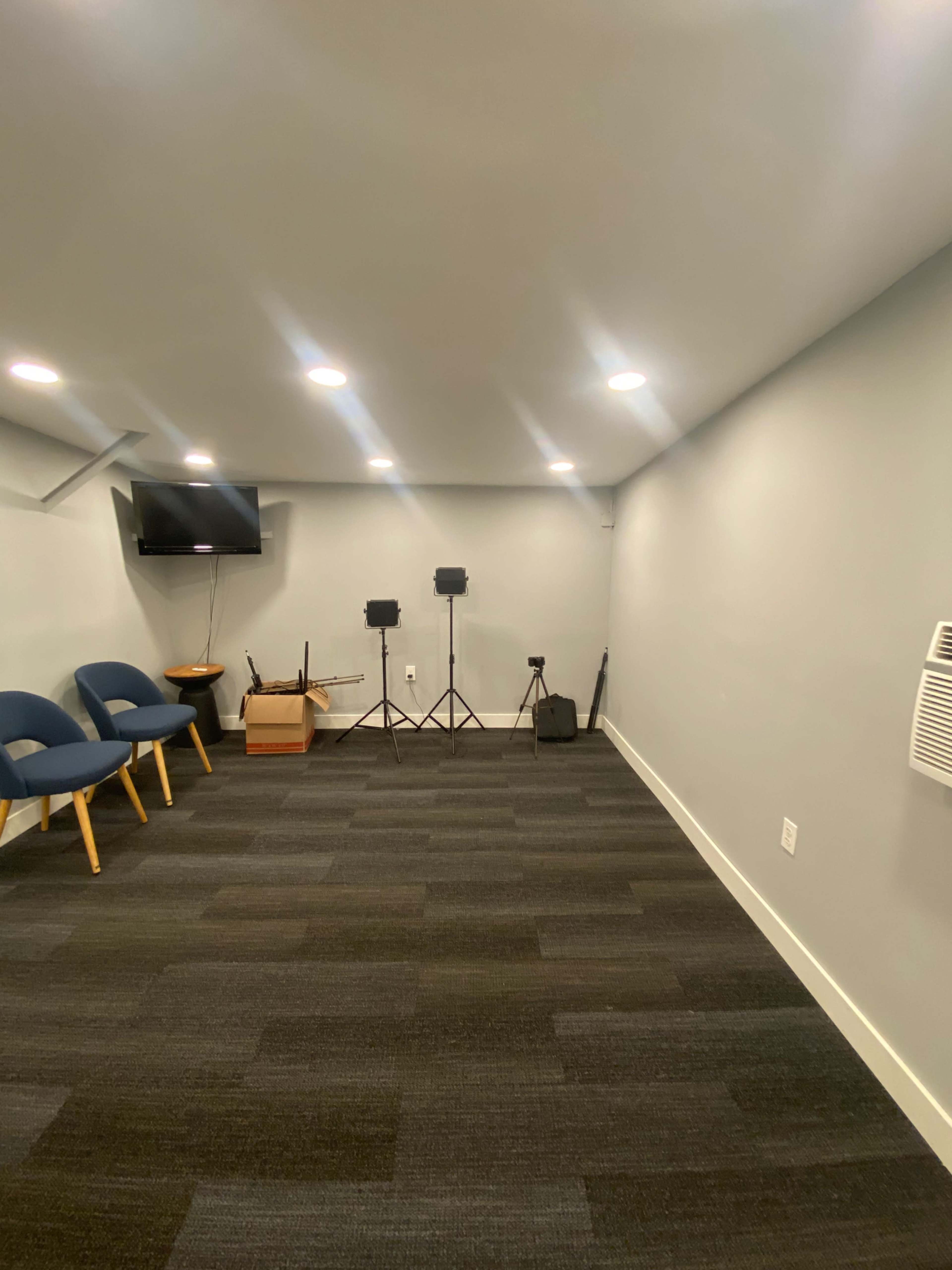 The image shows a sparsely furnished room with a television mounted on the wall, two microphone stands, and a few chairs along one side.