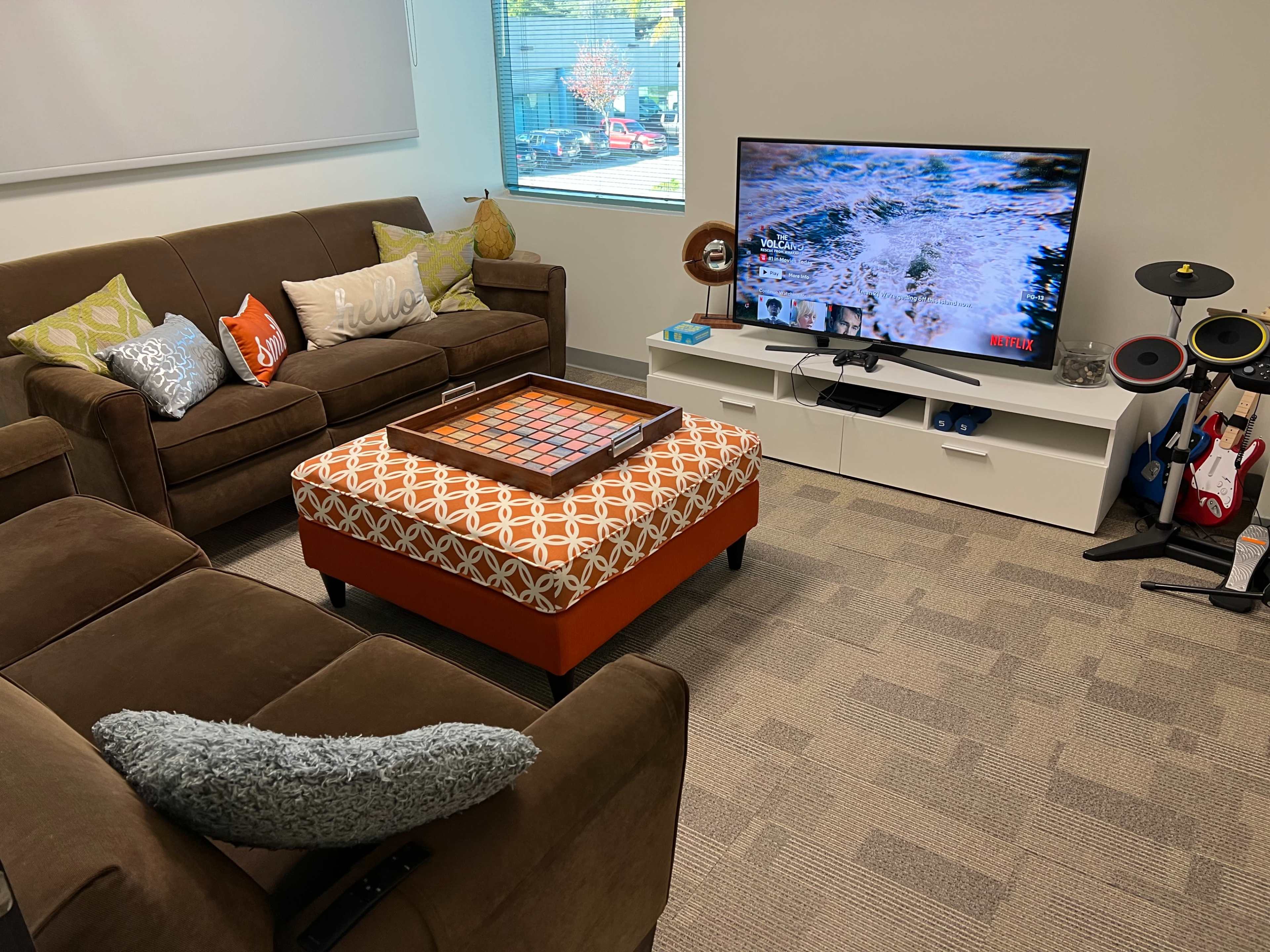 A cozy lounge area with a brown sofa set, a patterned ottoman, and a television on a white media console displaying content from Netflix.