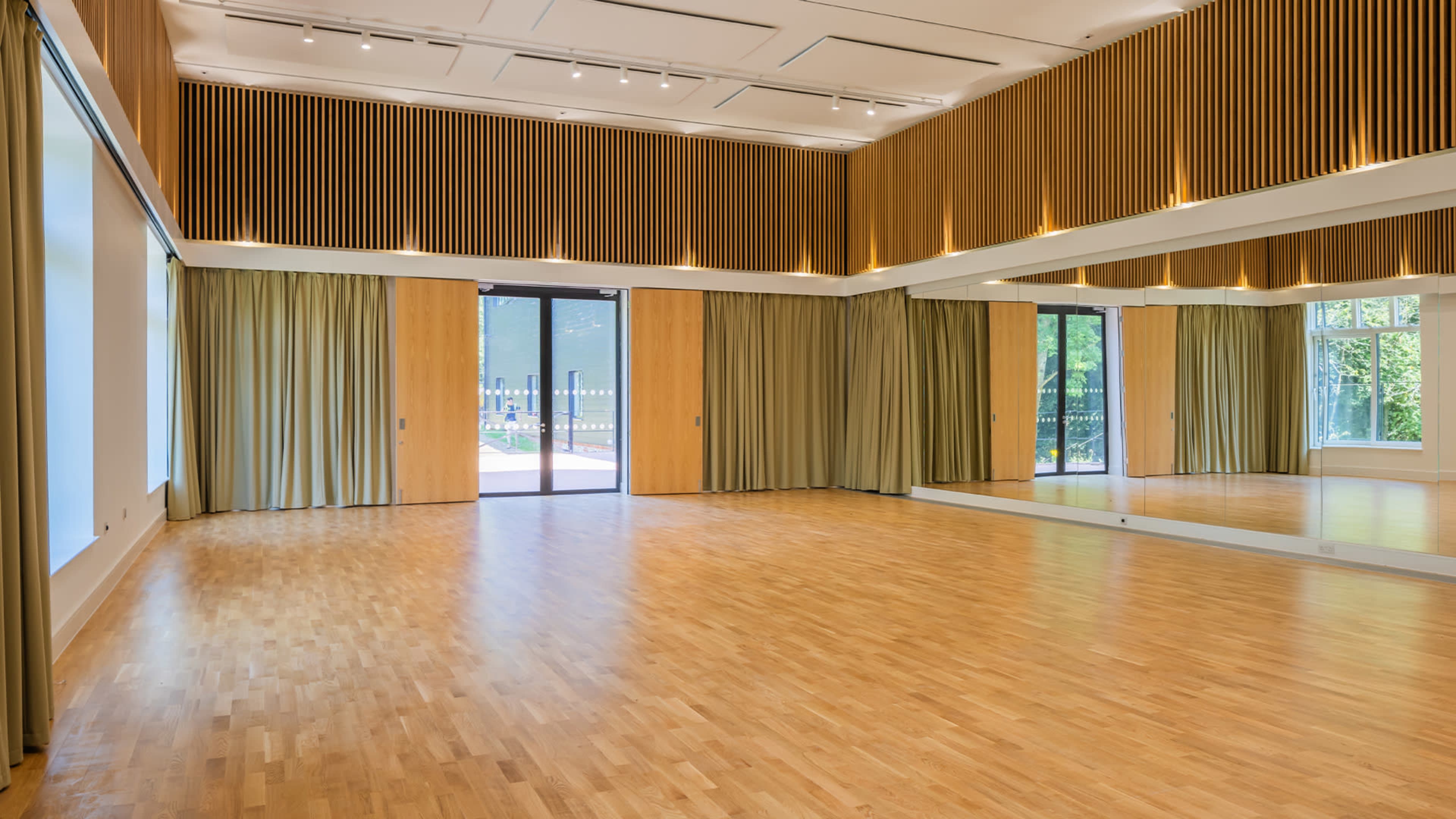 A spacious, empty interior room with wooden flooring, large windows, and curtains, along with a floor-to-ceiling mirror on one wall.