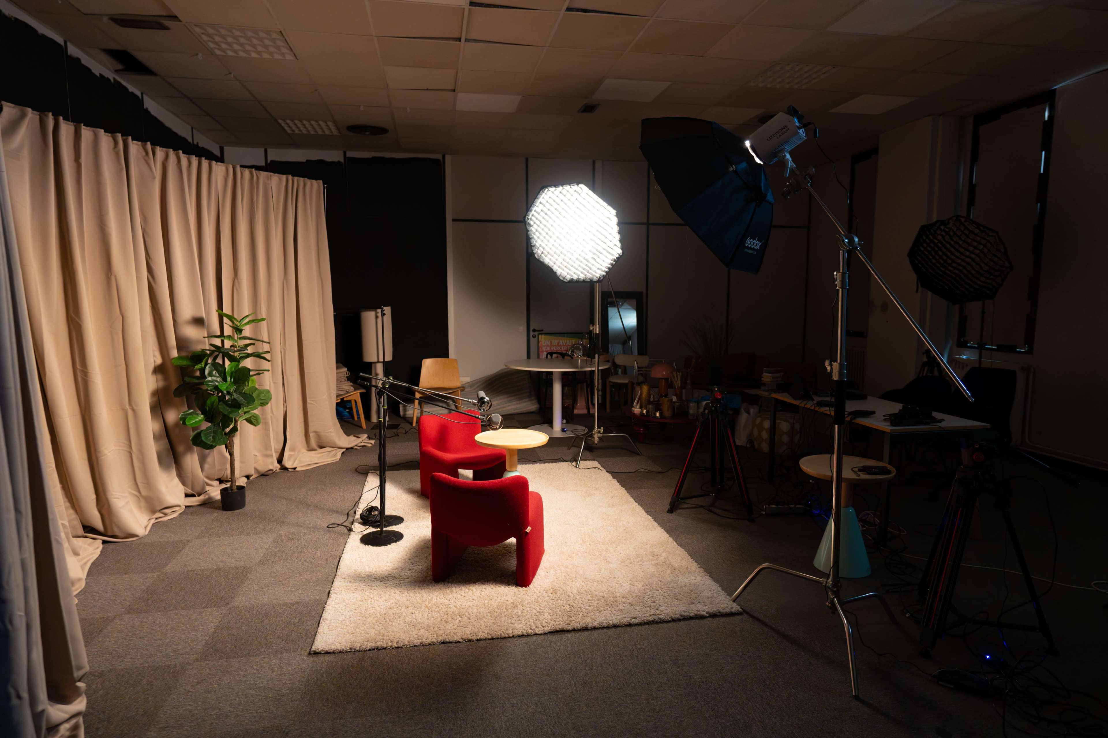 The image shows a well-lit studio setup featuring two red armchairs, a small round table, plants, and various lighting equipment.