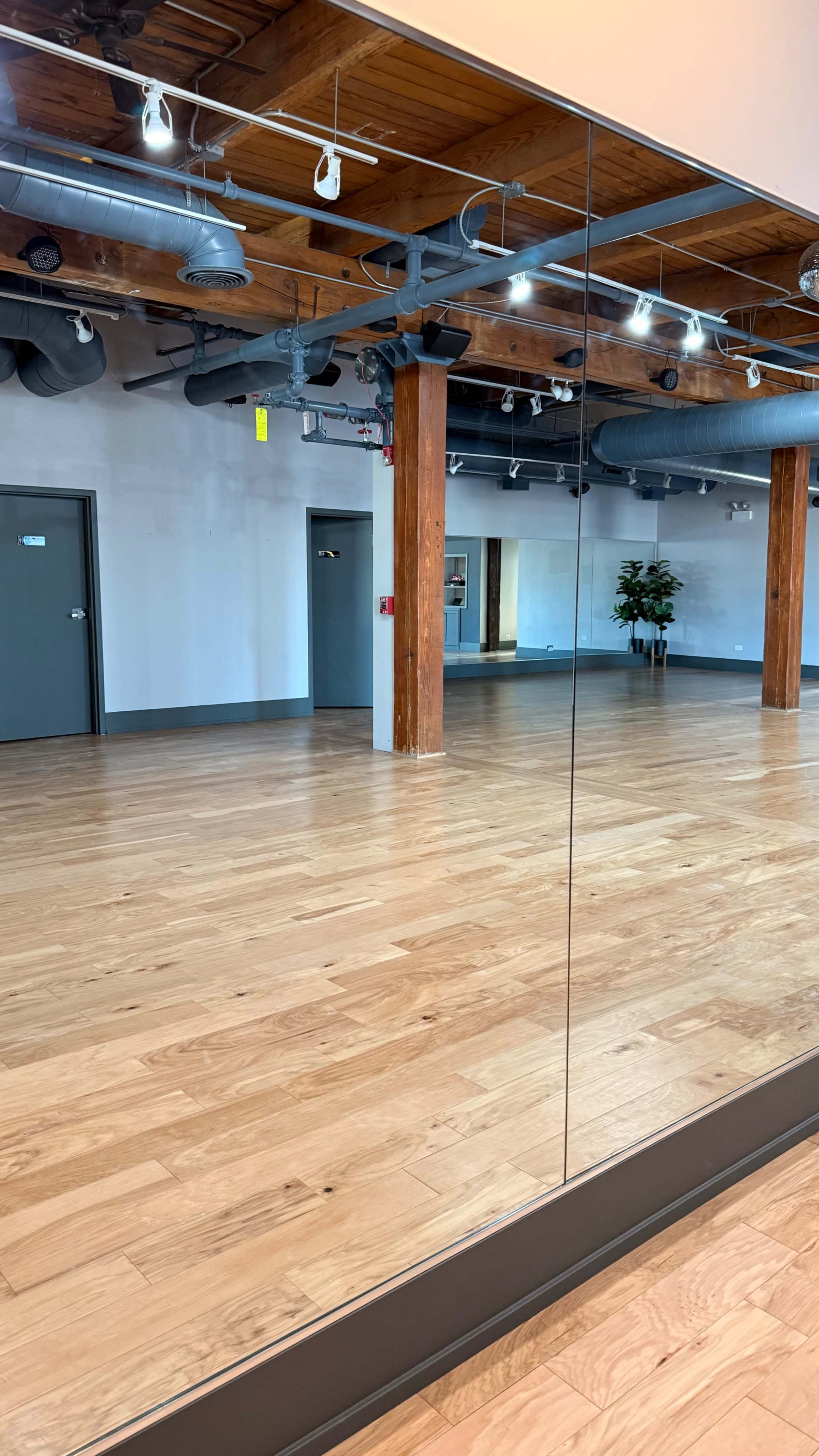 The image shows a spacious, empty room with wooden flooring, exposed wooden beams, and large mirrors on one wall.