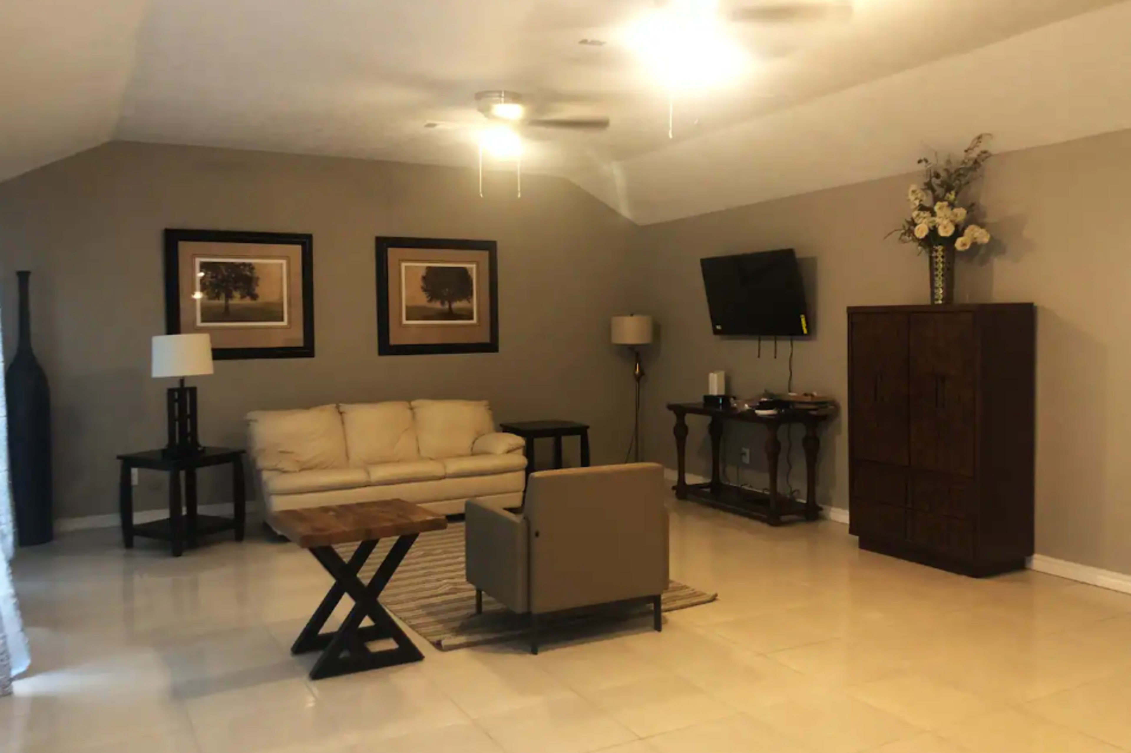 A living room with a beige couch, a small wooden coffee table, two table lamps, a television mounted on the wall, and a wooden cabinet.