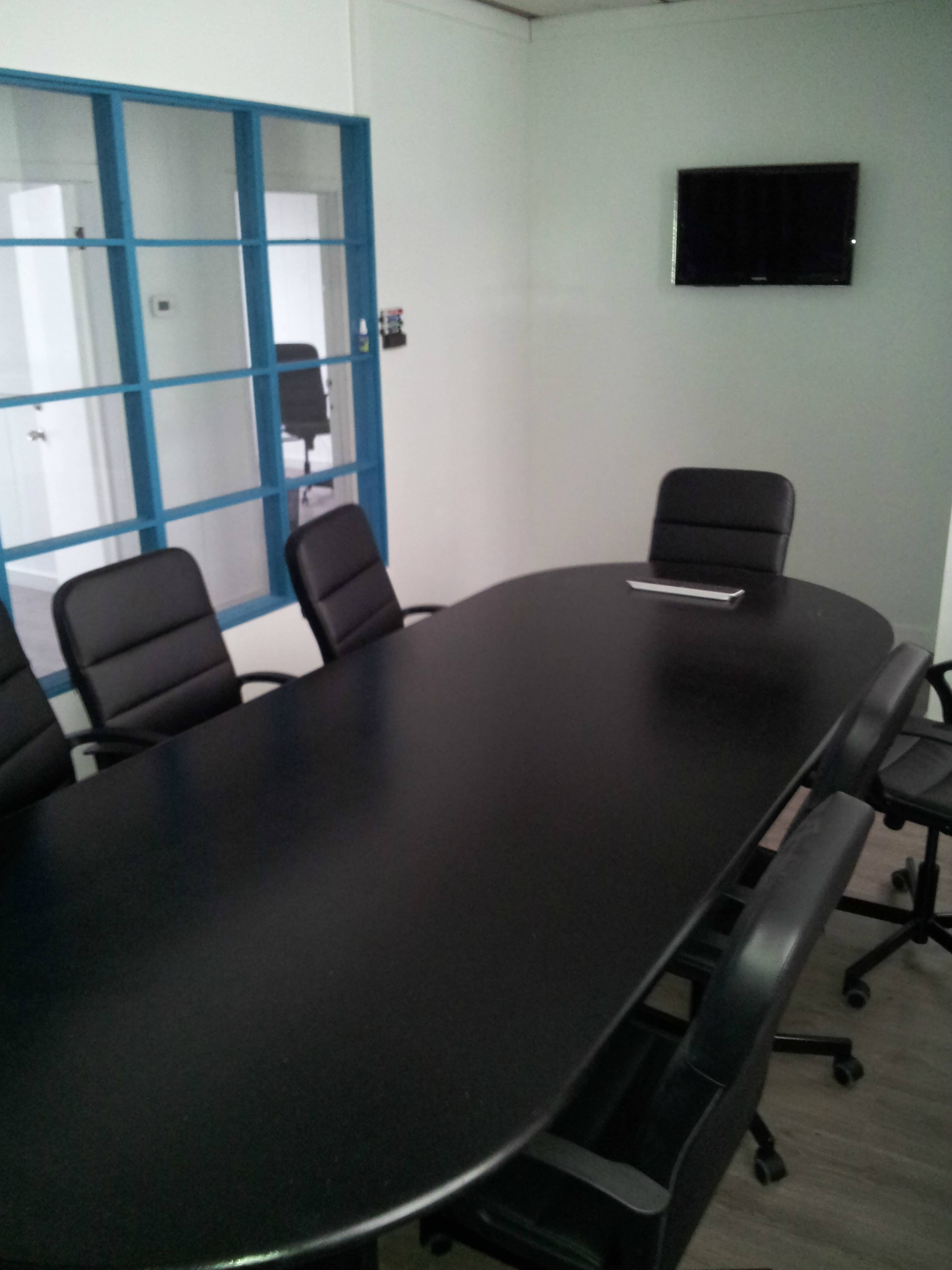 A conference room with a long black table surrounded by black rolling chairs, featuring a wall-mounted TV and a glass-paneled door.