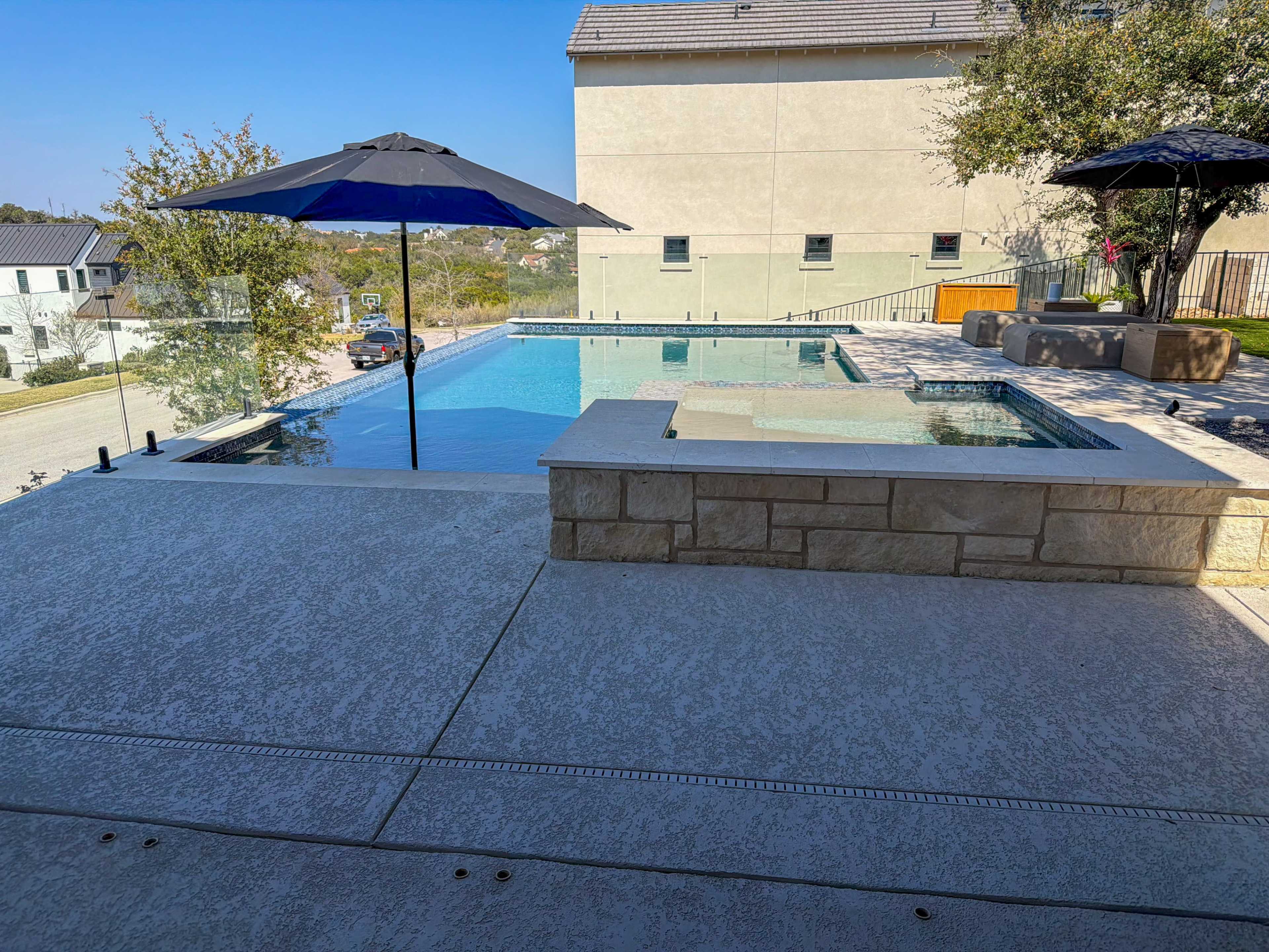 The image shows a pool with an infinity edge surrounded by a concrete deck, two umbrellas, and a view of houses in the distance.