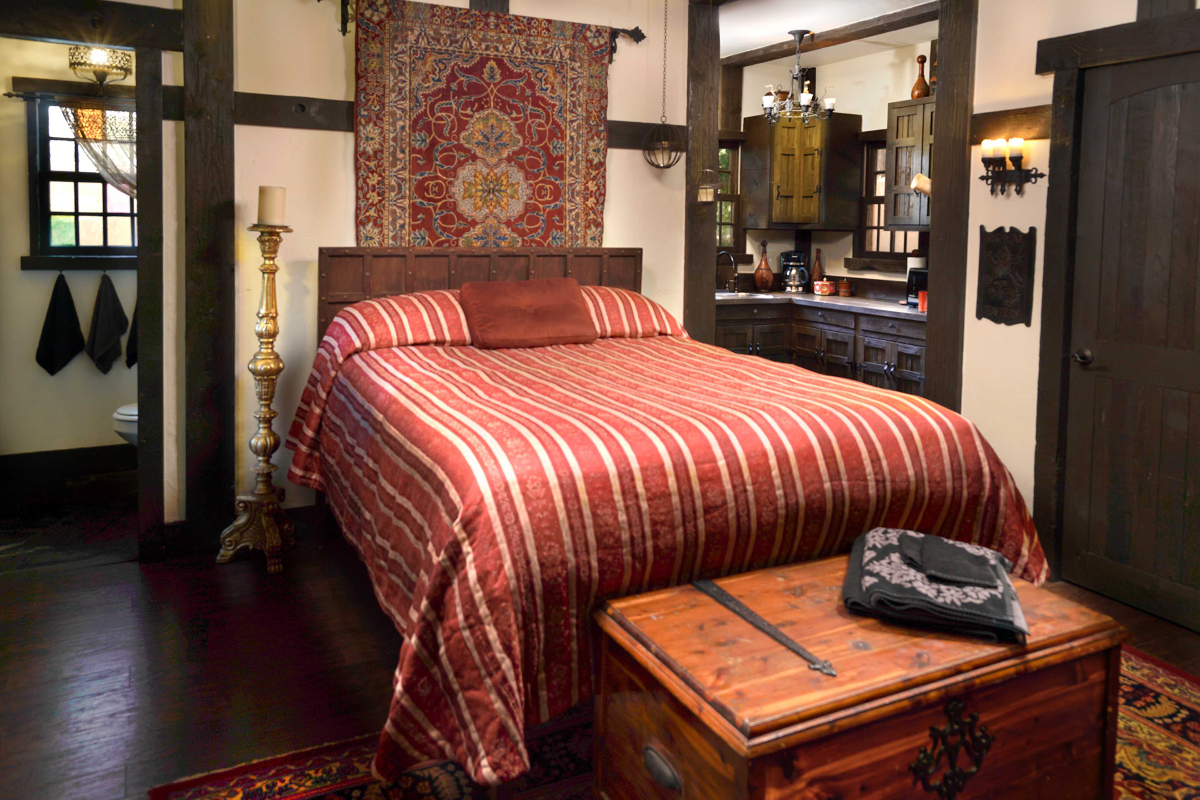 The image shows a bedroom with a striped red and gold bedspread, a wooden bed frame, a cozy chest at the foot of the bed, and a kitchenette visible in the background.