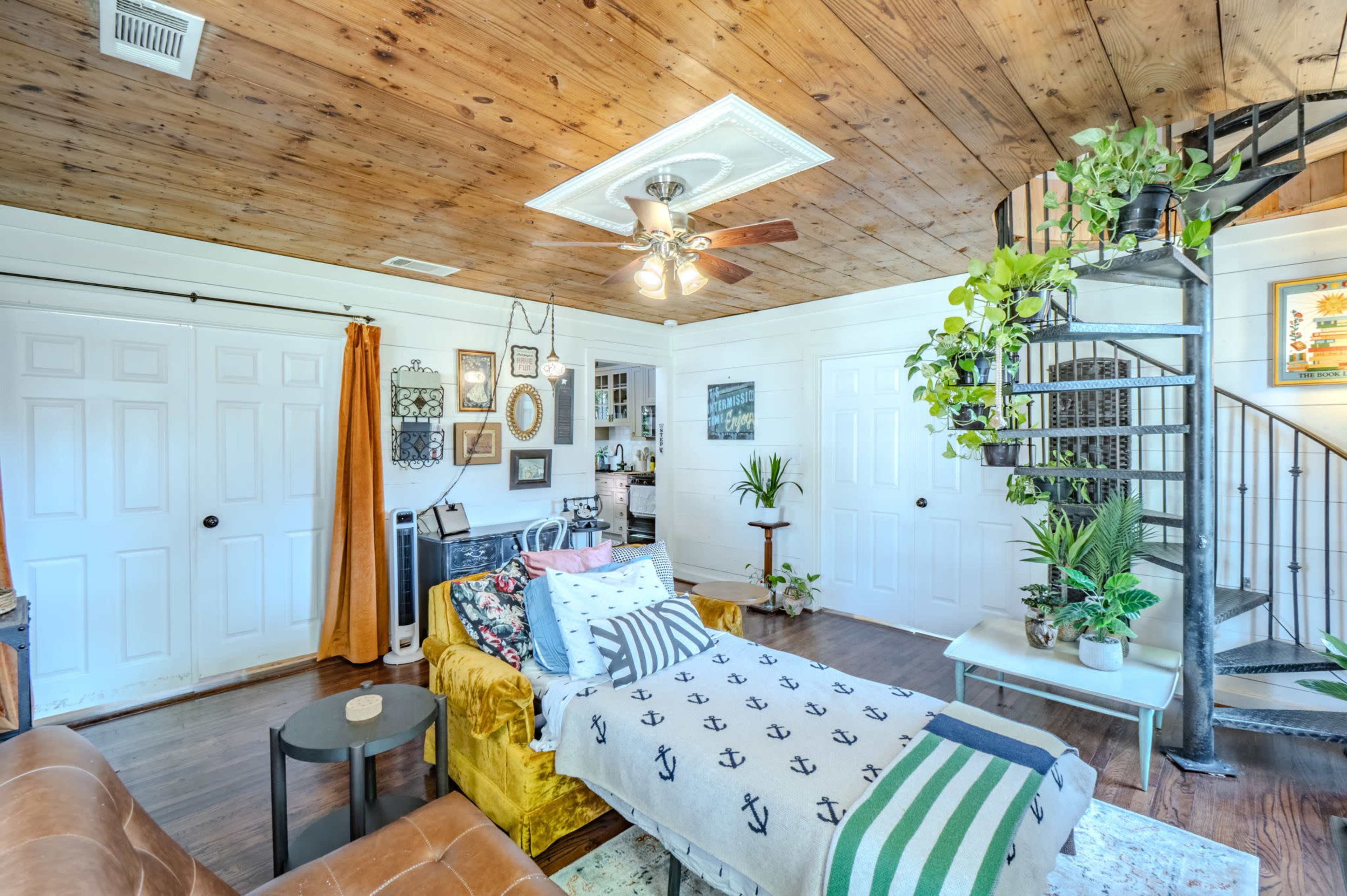 The living area features a yellow couch with patterned cushions, a spiral staircase, and decor including plants and framed artwork on the walls.