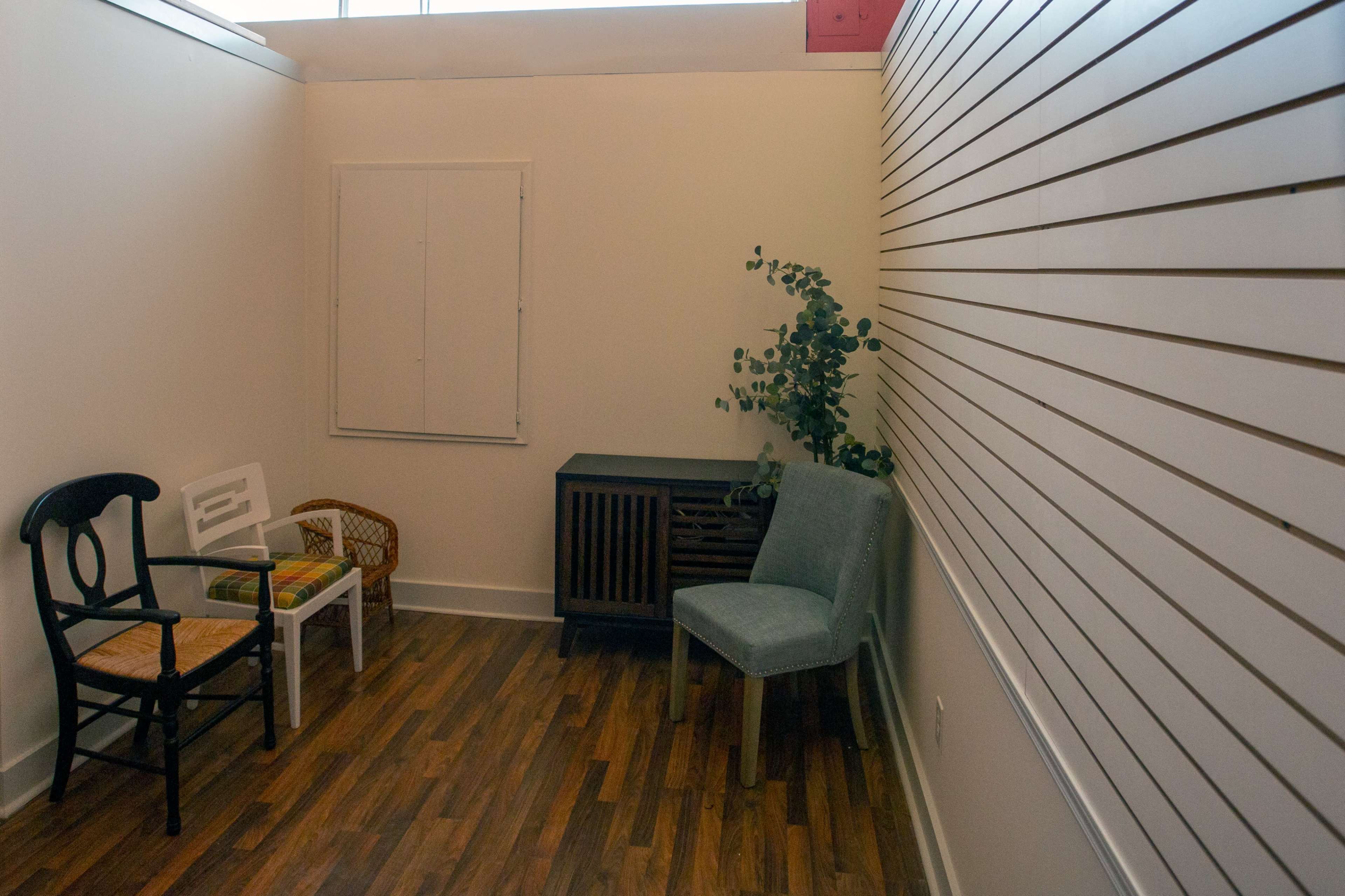 A small, brightly lit room features two chairs, a wooden cabinet, and a potted plant against a slatted wall.