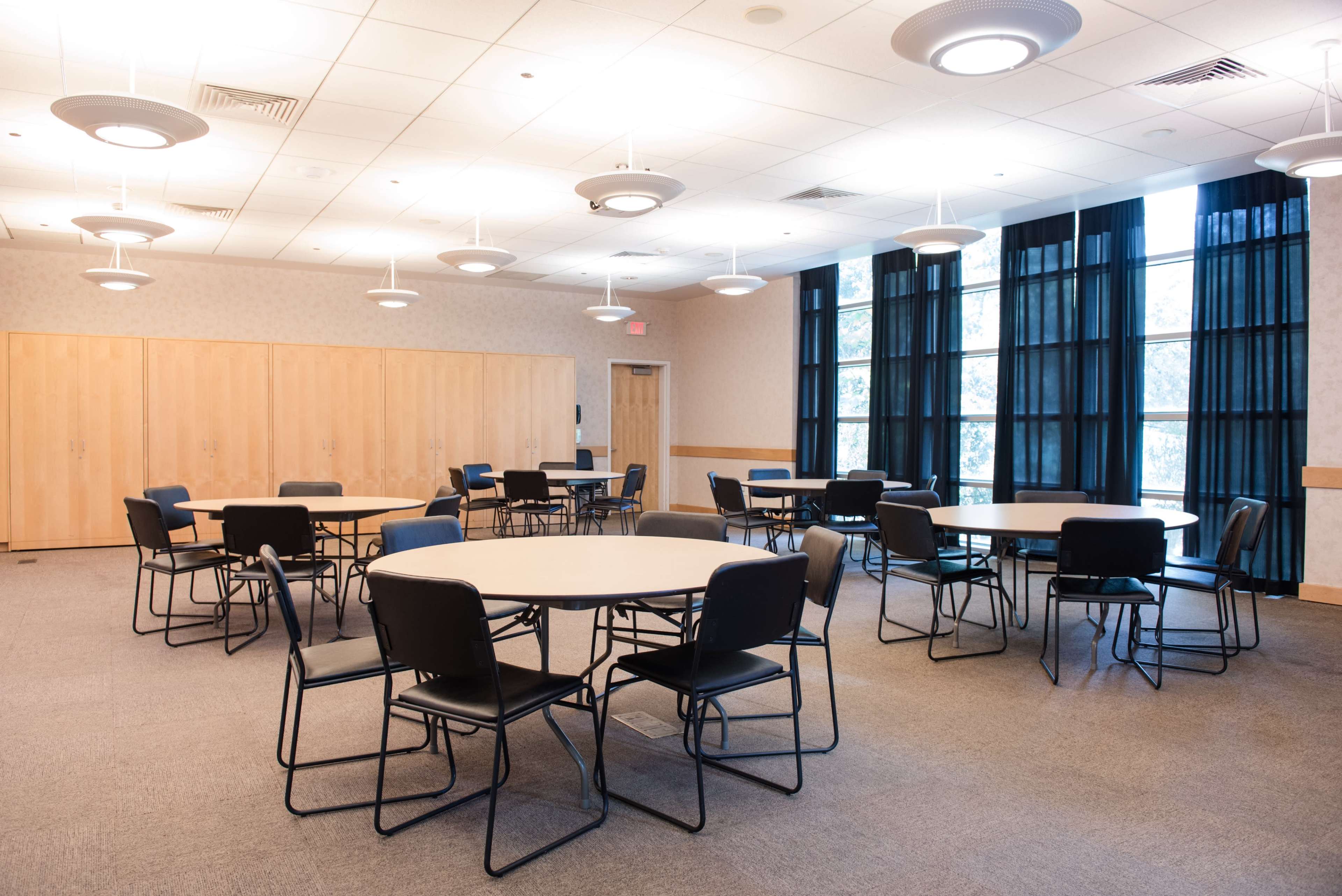 A meeting room features several round tables surrounded by black chairs, with large windows and suspended lights above.