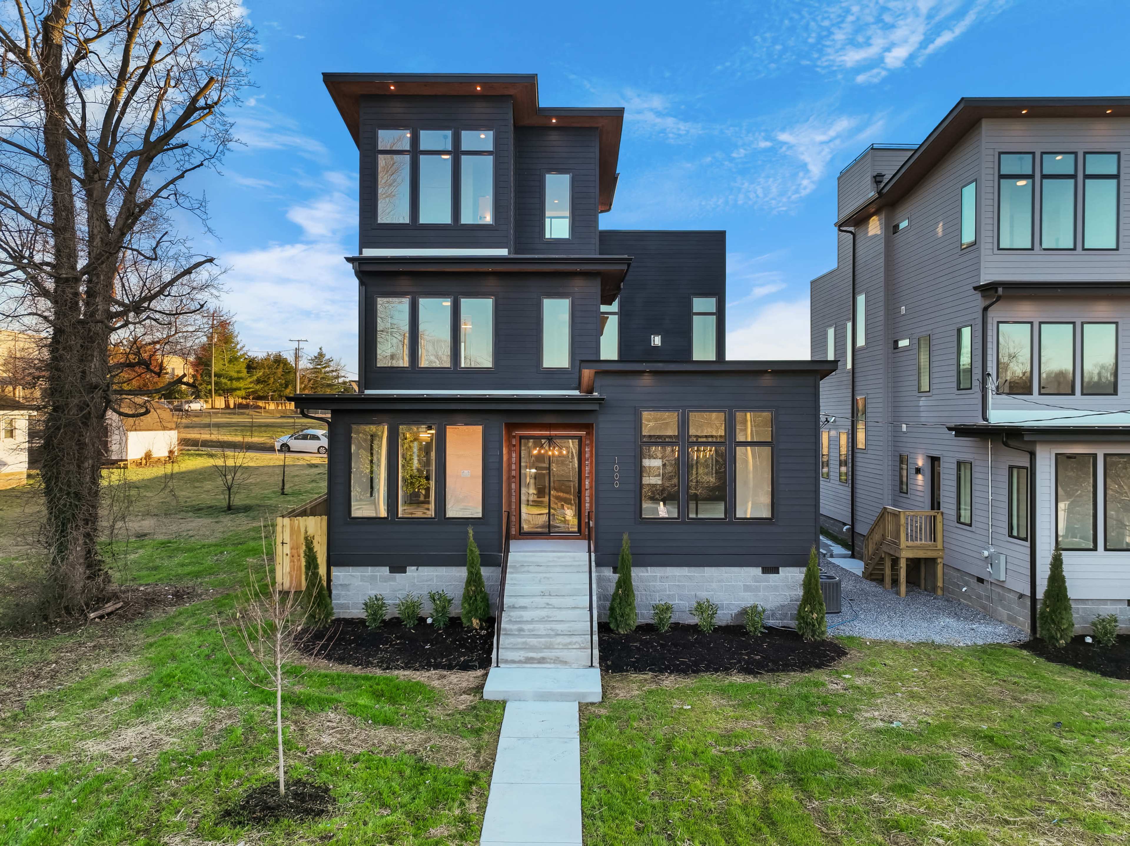 The image shows a modern three-story house with large windows and a landscaped front yard, situated beside another contemporary building.