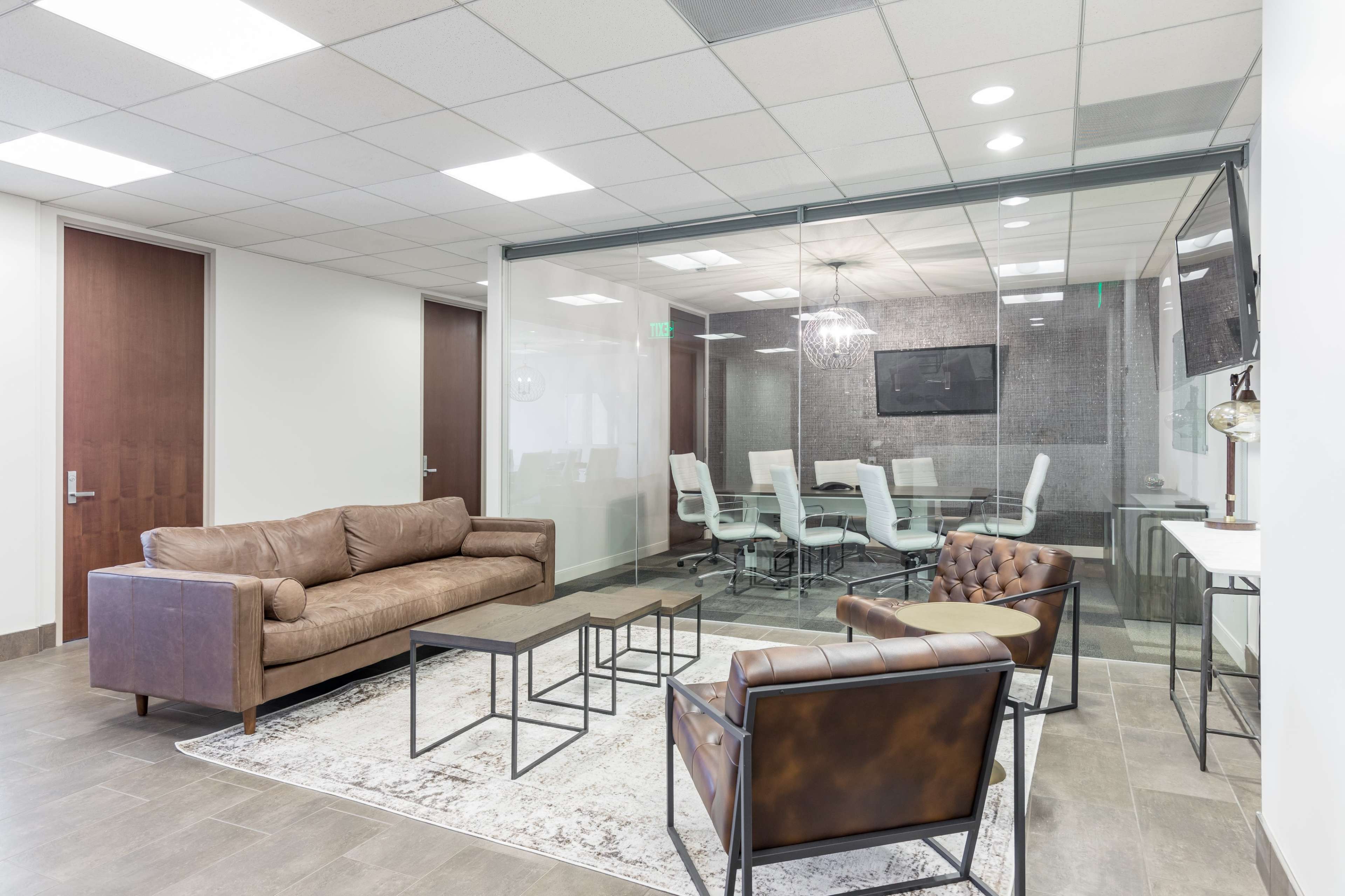 A modern office lounge area features a brown leather sofa, two armchairs, a coffee table, and a glass-walled meeting room with white chairs and a television.