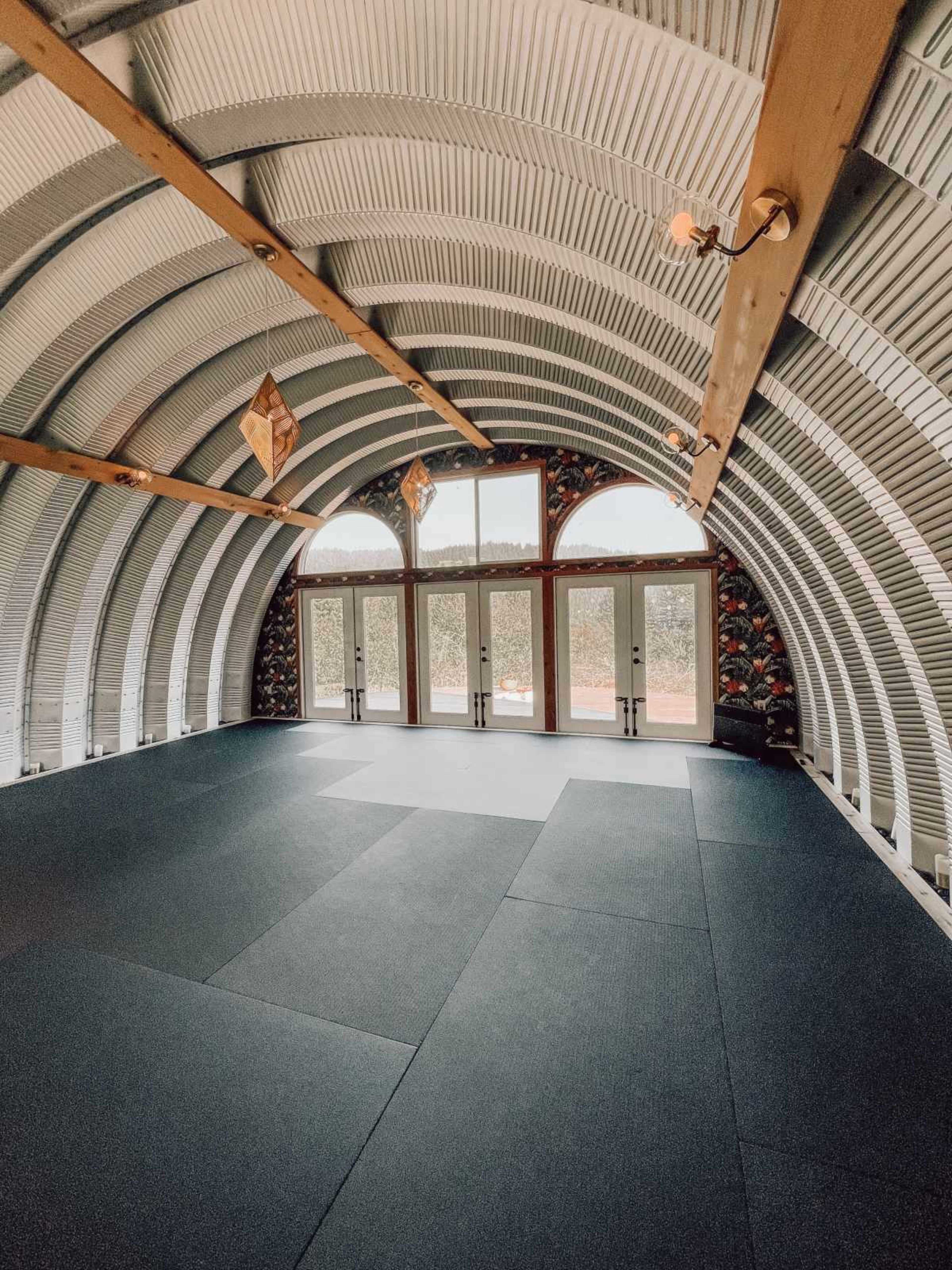 The image shows the interior of a large, arched metal building with a padded floor and glass doors leading outside.