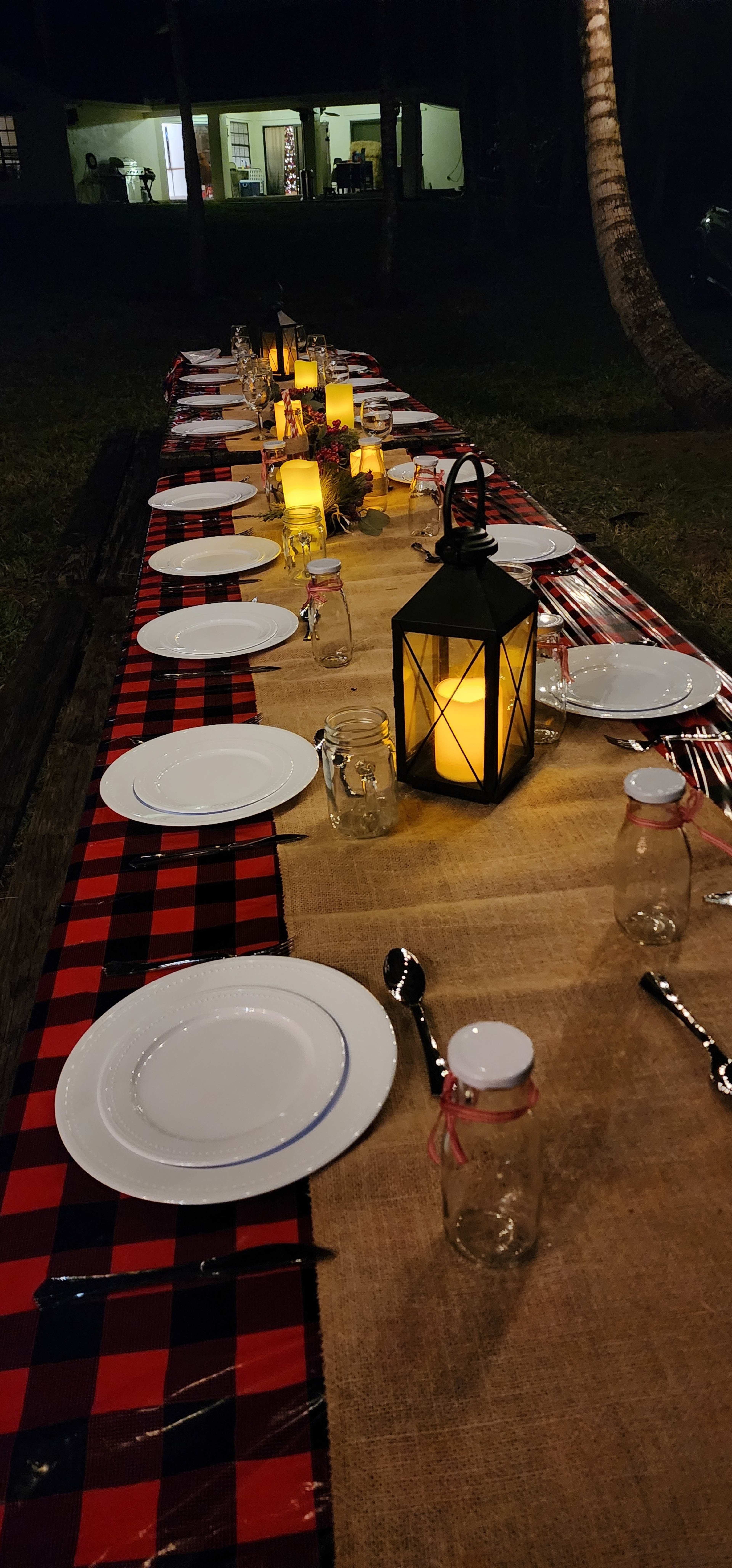 A long outdoor dining table is set with white plates, mason jars, and a lantern, all arranged on a red and black checkered tablecloth.