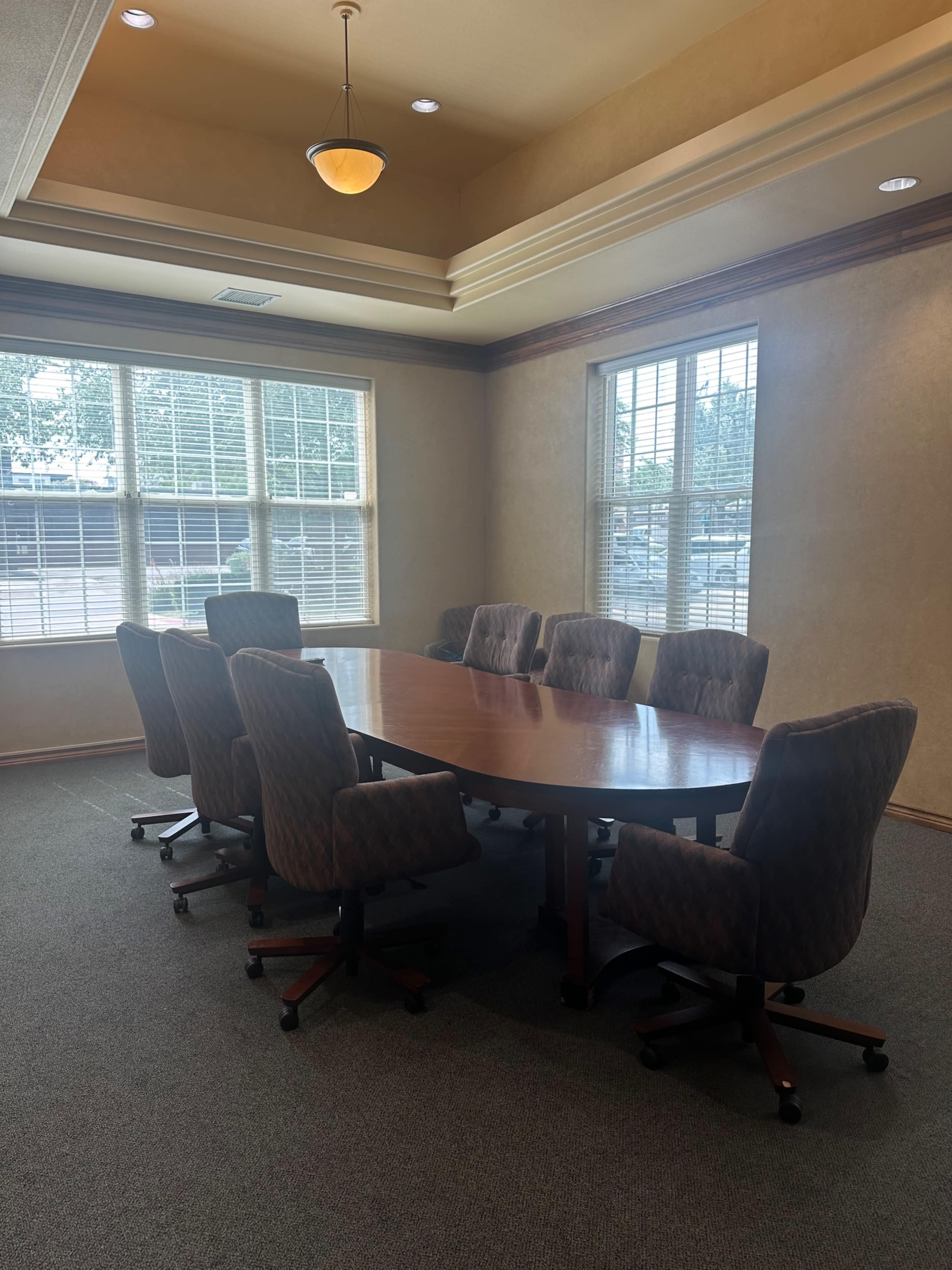 The image shows a conference room with a large wooden table surrounded by eight upholstered chairs and bright windows allowing natural light.