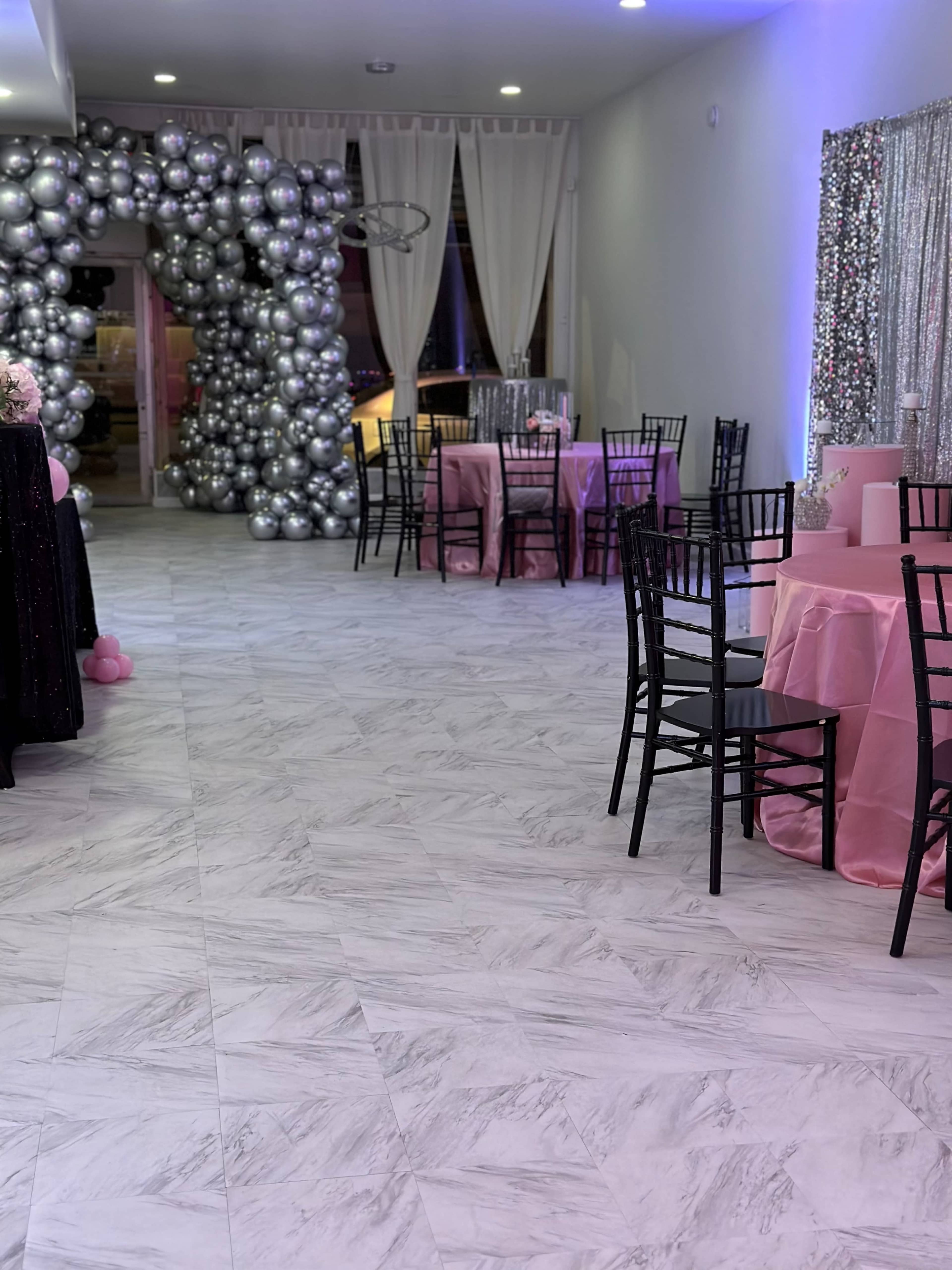A decorated event space with black chairs and pink tables, accented by a large balloon arch and shimmering curtains.