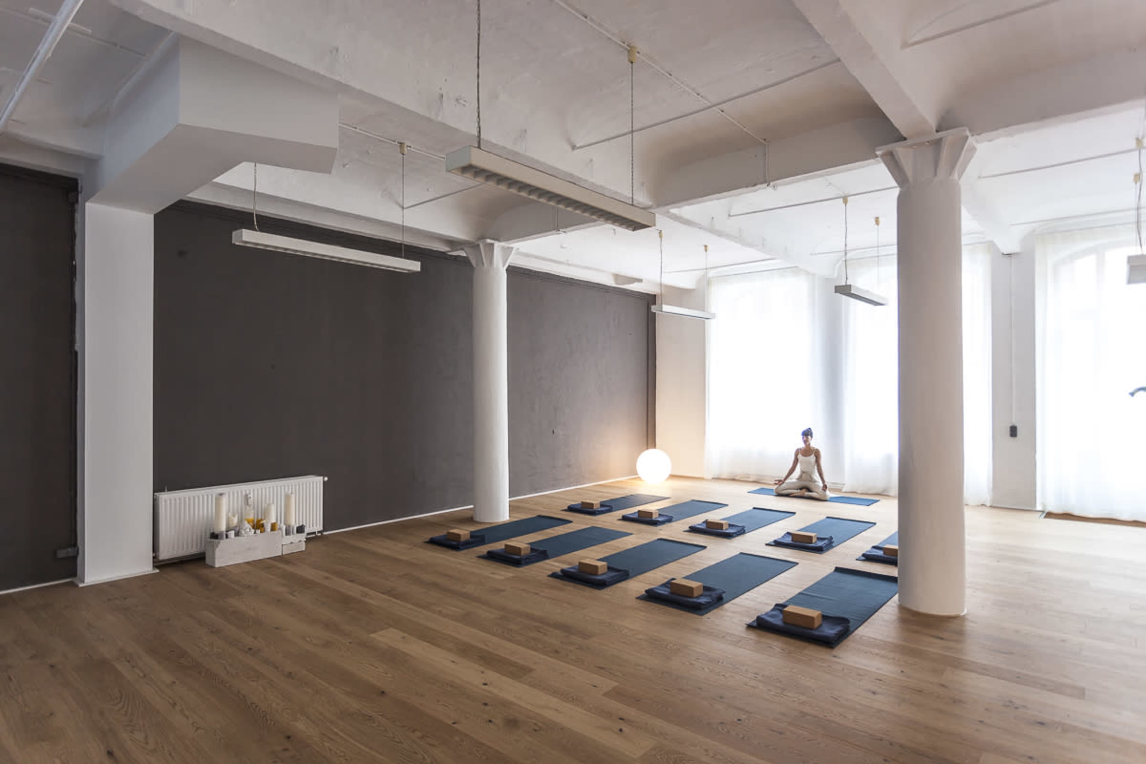 A spacious yoga studio with wooden flooring, white pillars, and exercise mats arranged neatly on the floor.