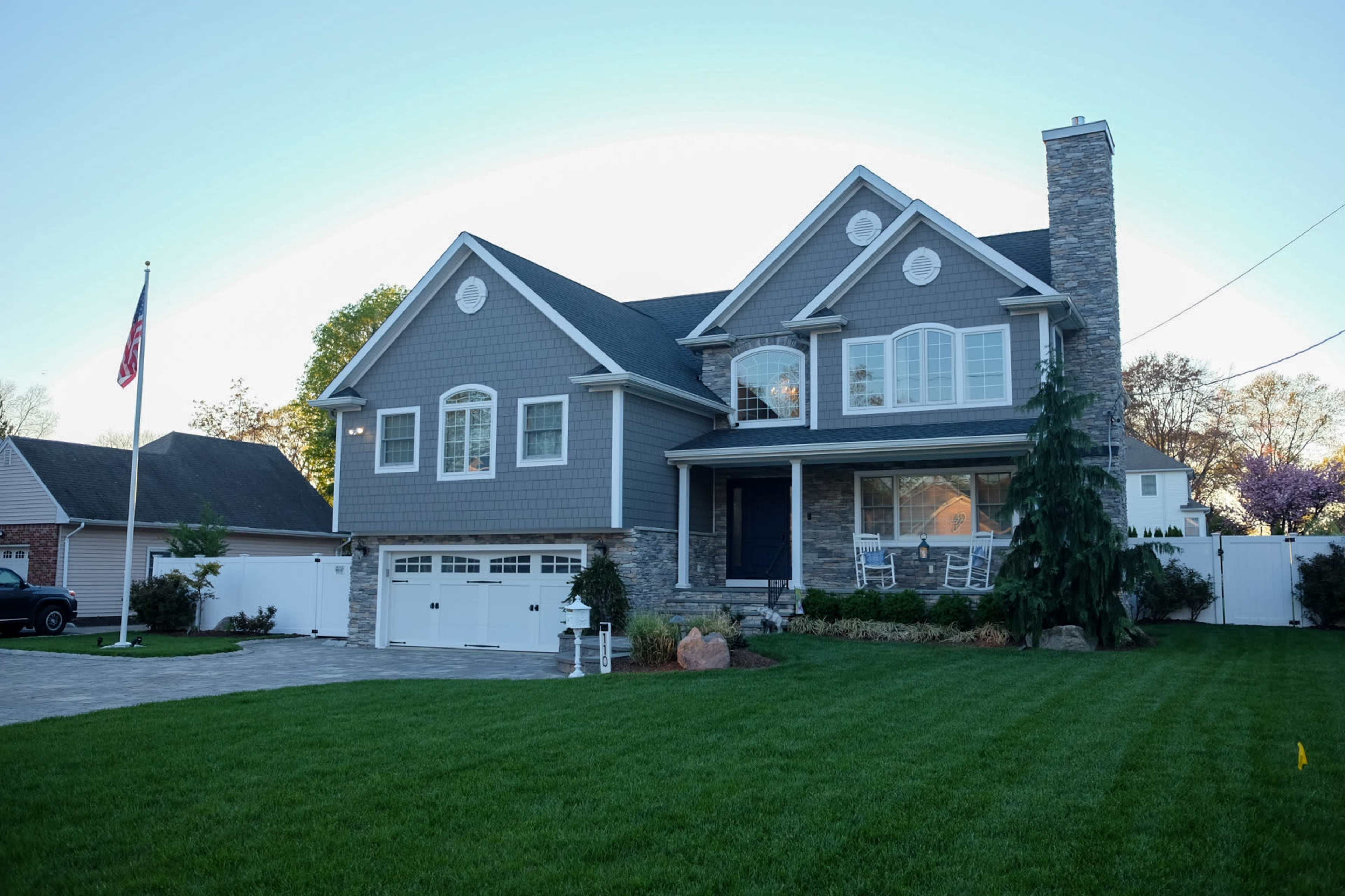 A two-story gray house with stone accents and a front porch, flanked by a neatly landscaped yard and an American flag.
