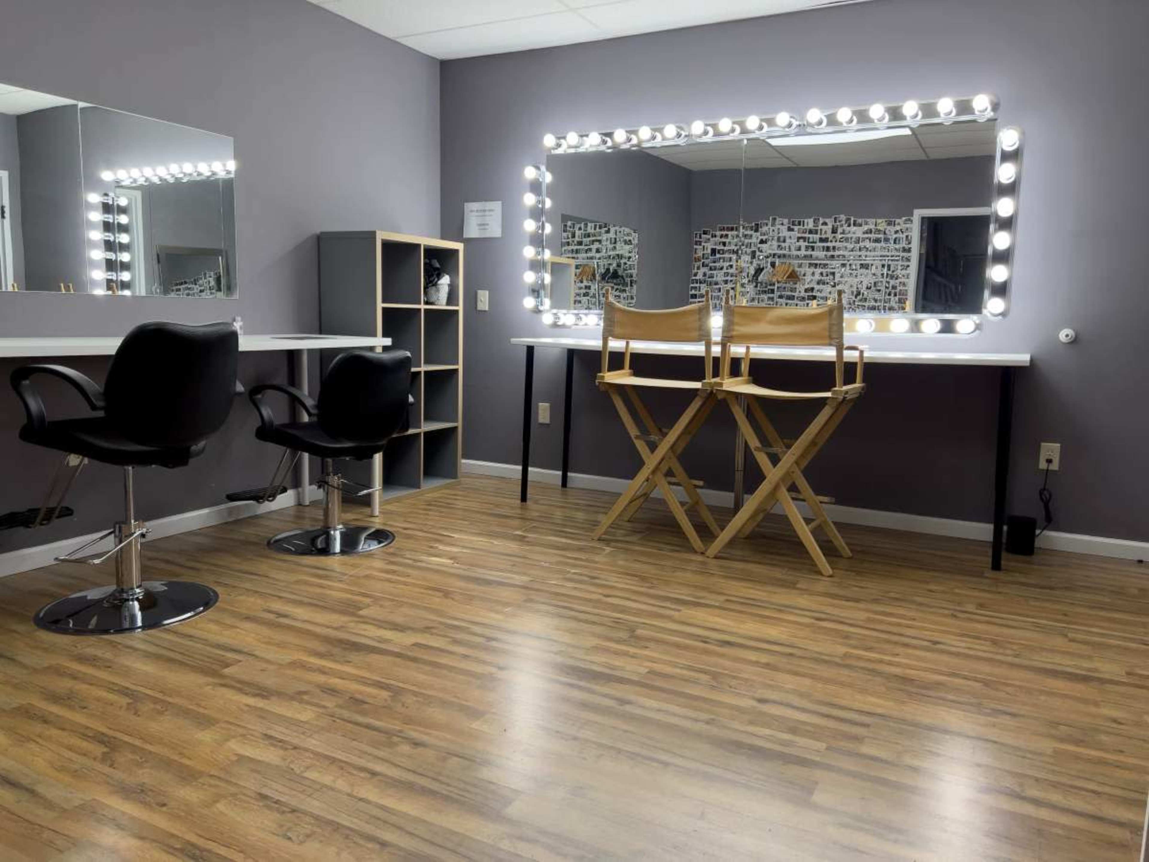 The image shows a spacious room with two black salon chairs, a mirrored vanity with bright lights, and a folding chair set against a gray wall.