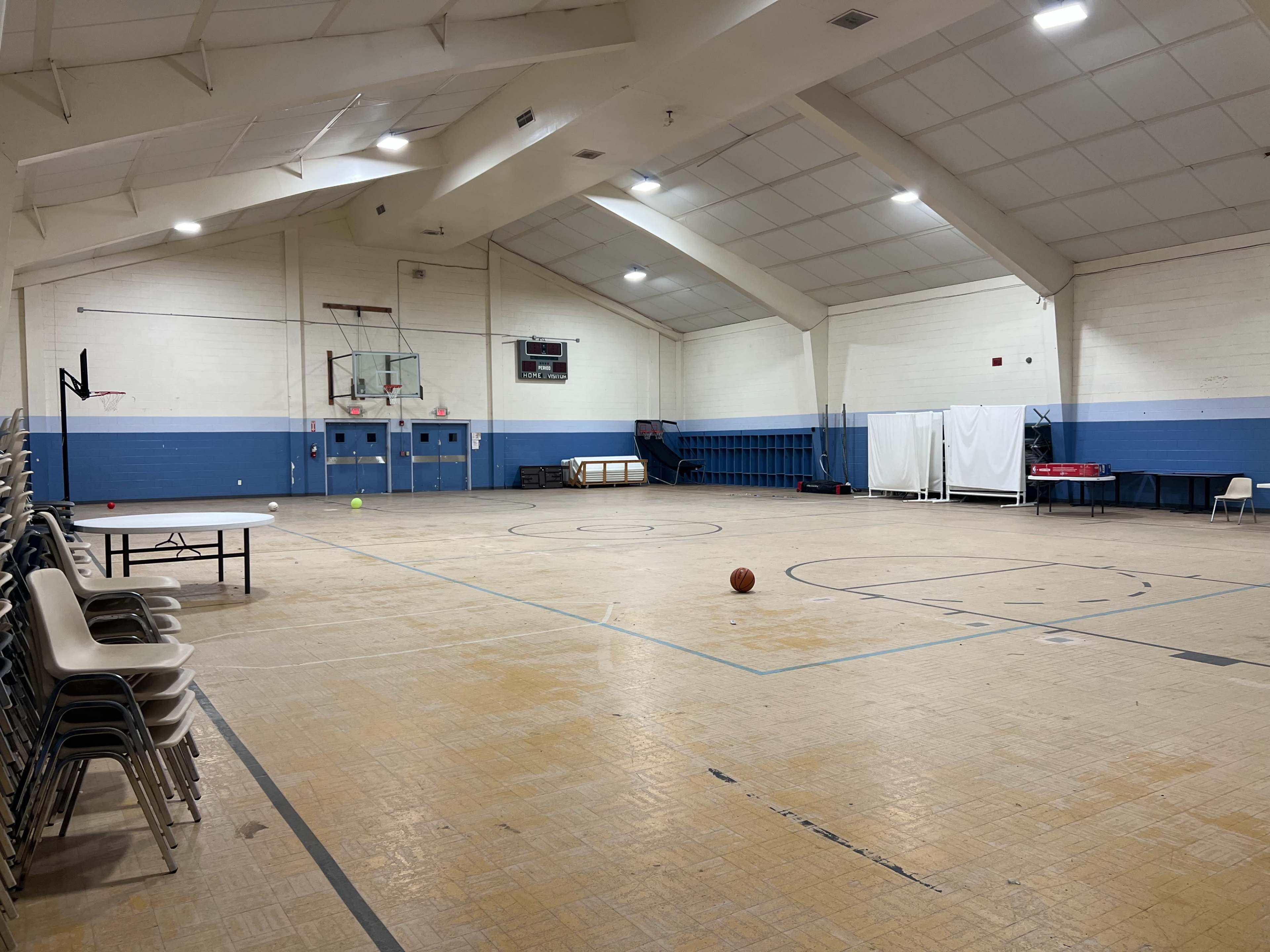 The image shows a large, empty indoor gymnasium with basketball hoops, a few scattered balls, and folding chairs stacked against one wall.