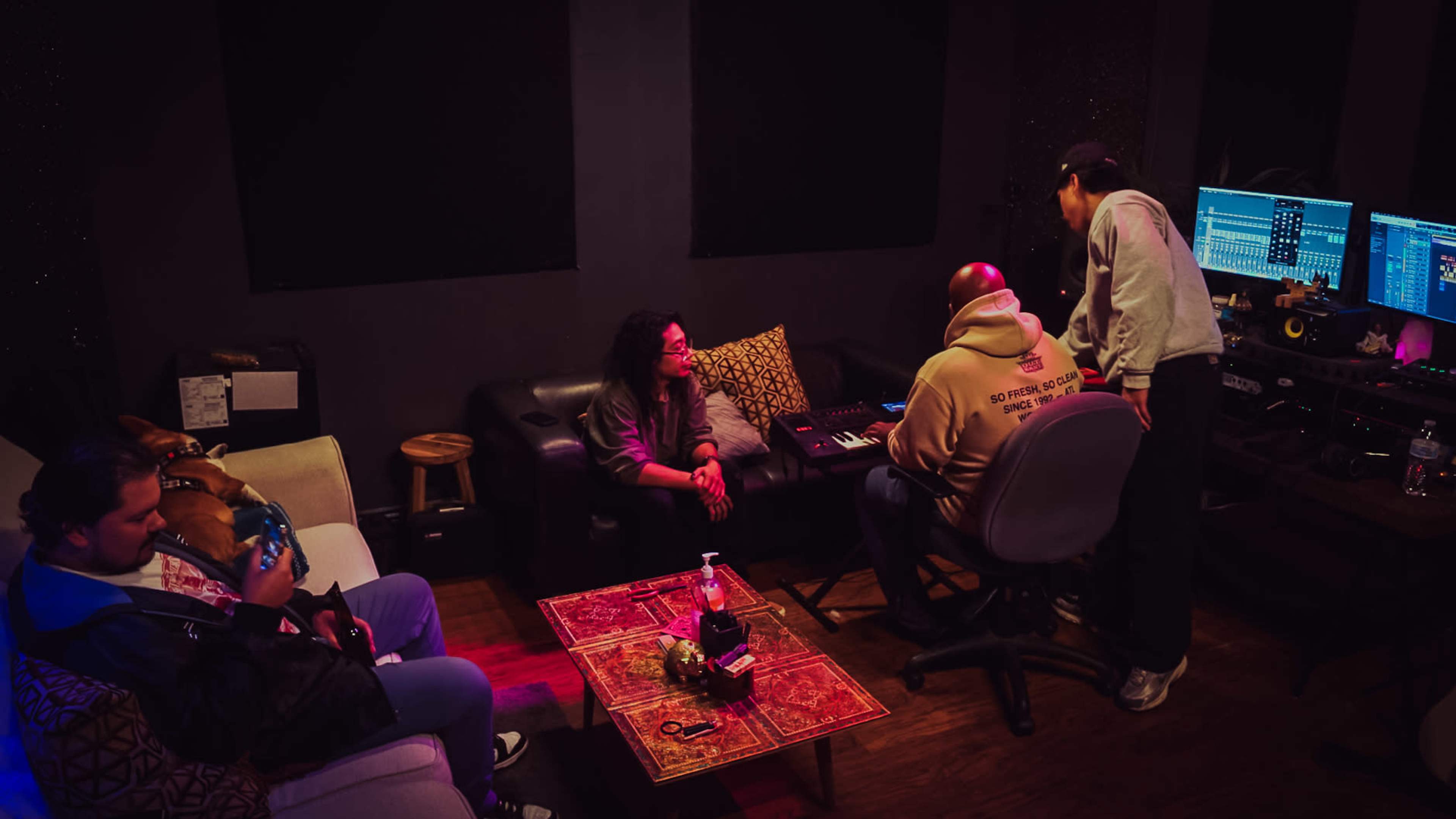 A group of four people is collaborating in a music studio filled with equipment and dim lighting.
