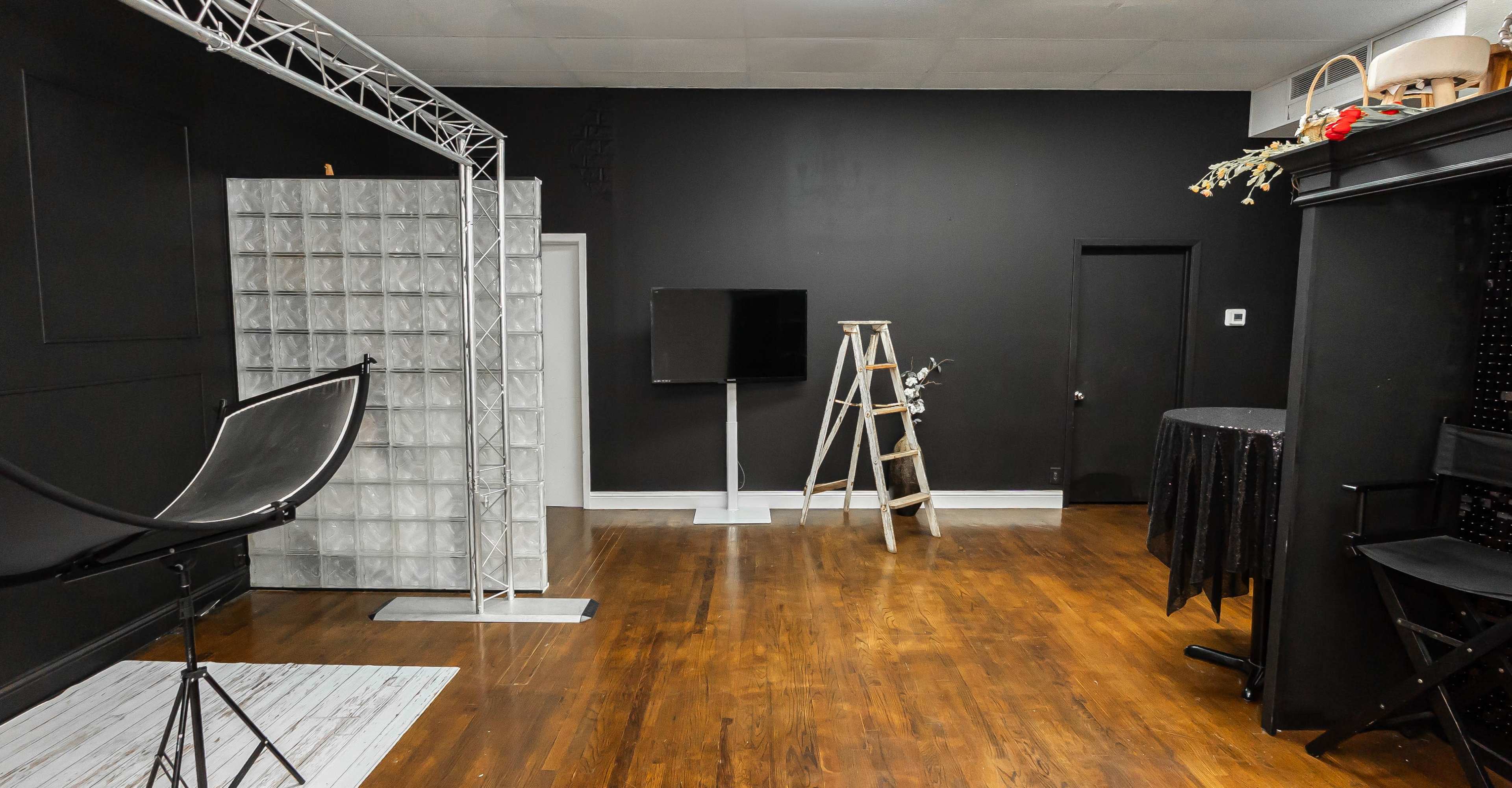 The image depicts an empty studio space featuring a black wall, a glass block partition, a television on a stand, a ladder, and various furniture pieces.