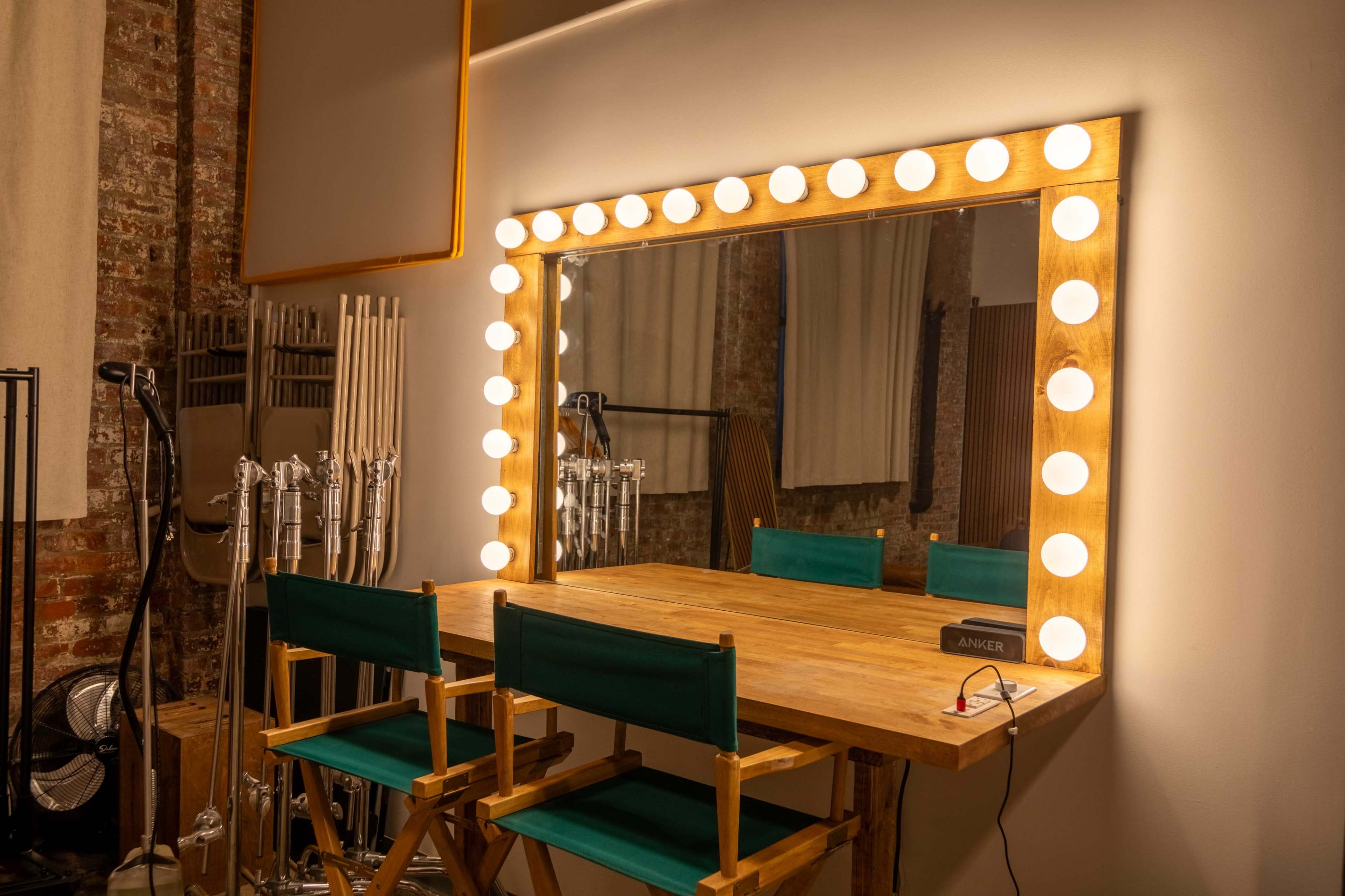 A brightly lit makeup mirror with a wooden frame and several makeup chairs is positioned against a brick wall.