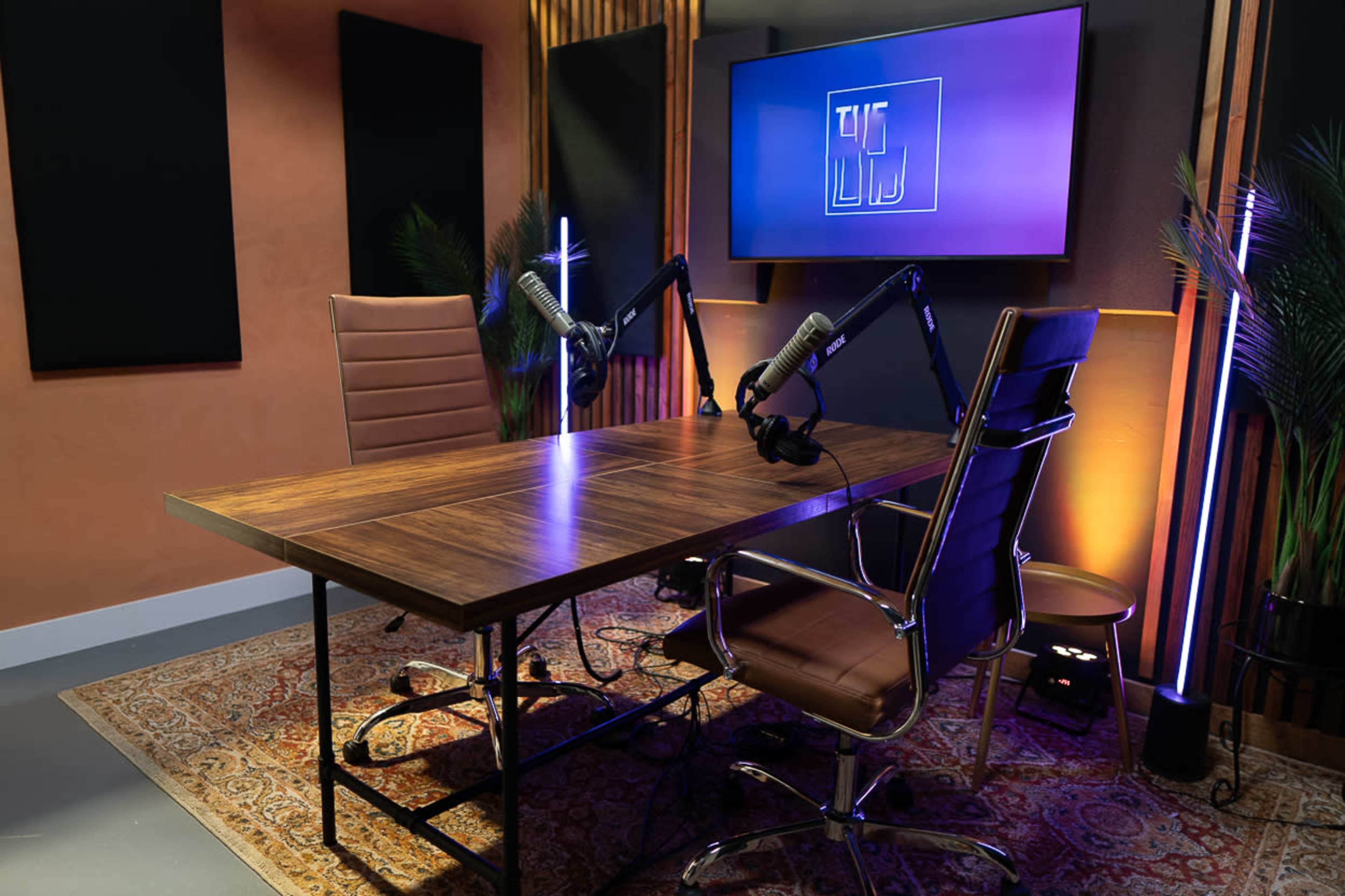A modern podcast studio features a wooden table, two chairs, and microphones mounted on stands, with ambient lighting and a large screen displaying a logo.