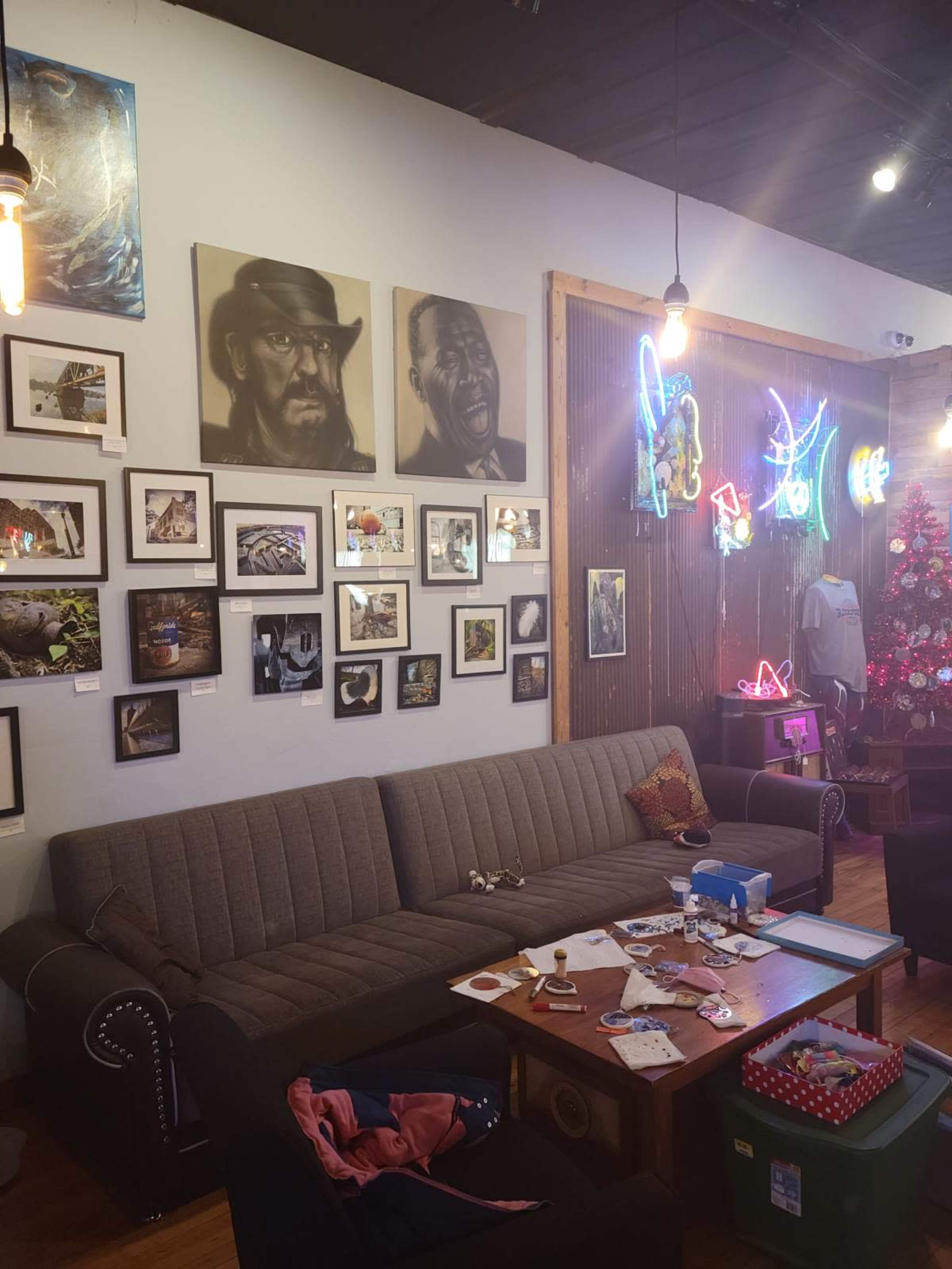 The image shows an interior of a cafe or gallery with a gray couch, a wooden table, various framed artworks on the walls, and colorful neon signs.