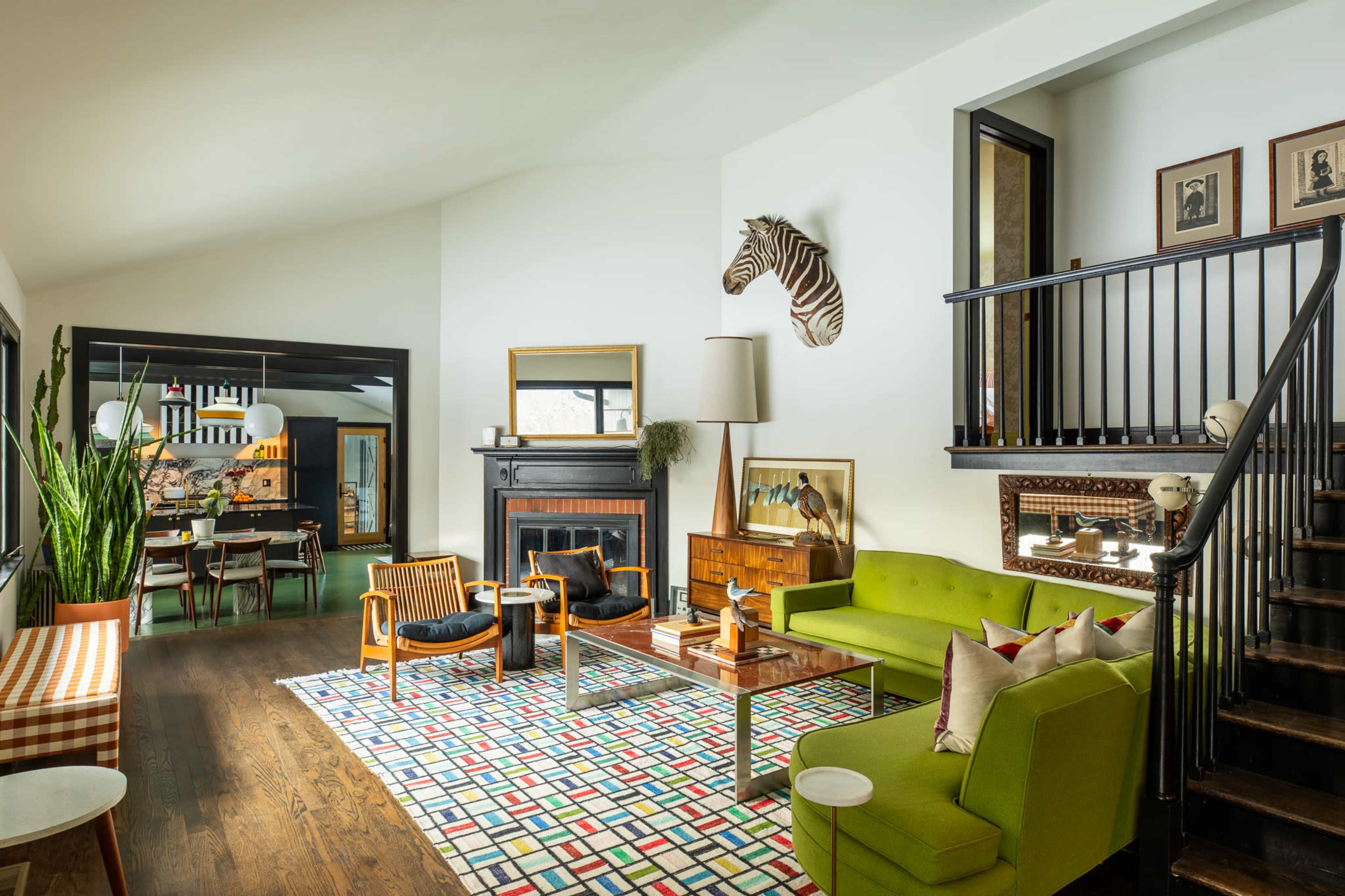 The image shows a contemporary living room with a mix of seating arrangements, a patterned rug, and a staircase leading to an upper level.