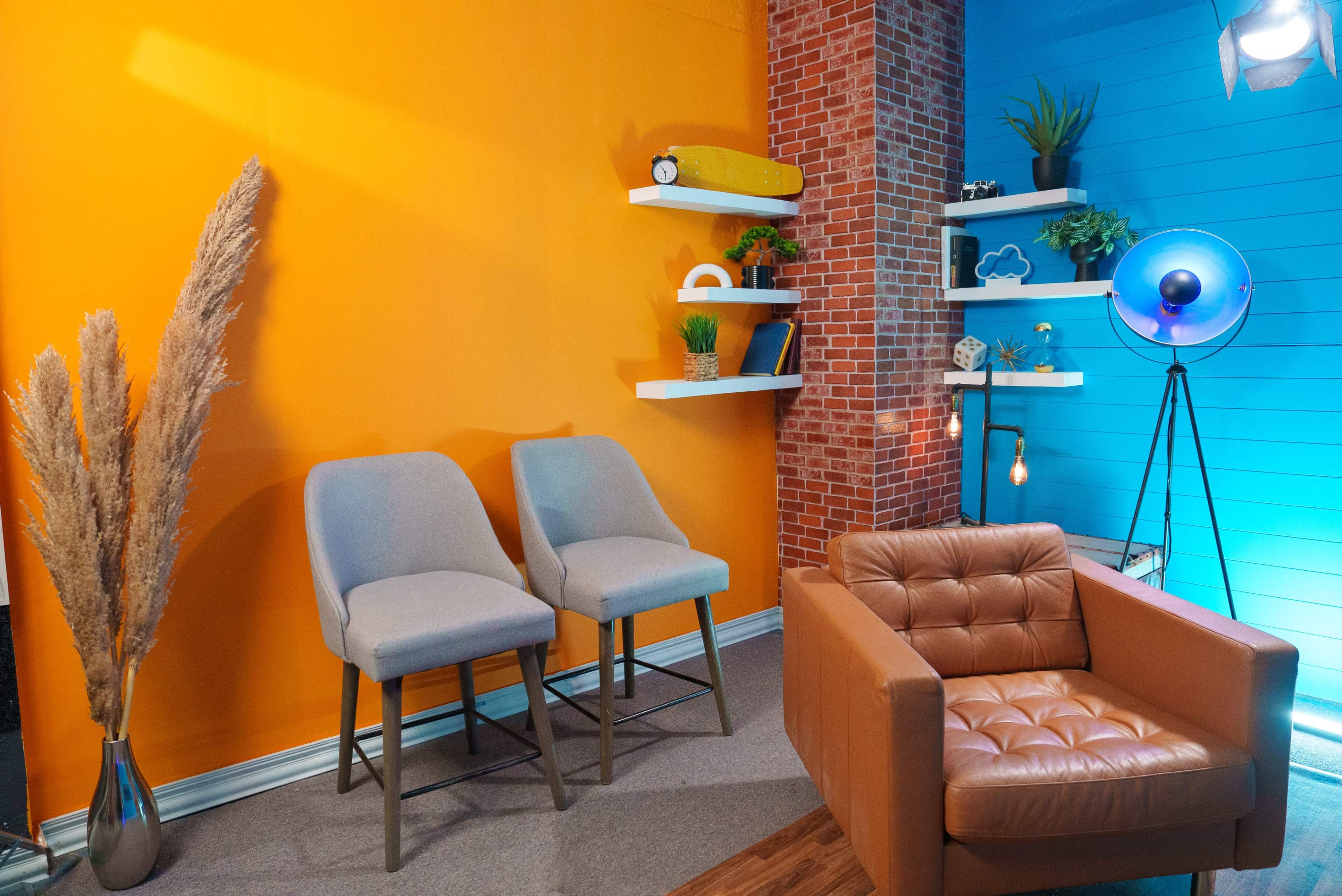 The scene features a modern interior with an orange and blue color scheme, showcasing two gray chairs, a brown leather armchair, decorative shelves, and plants.