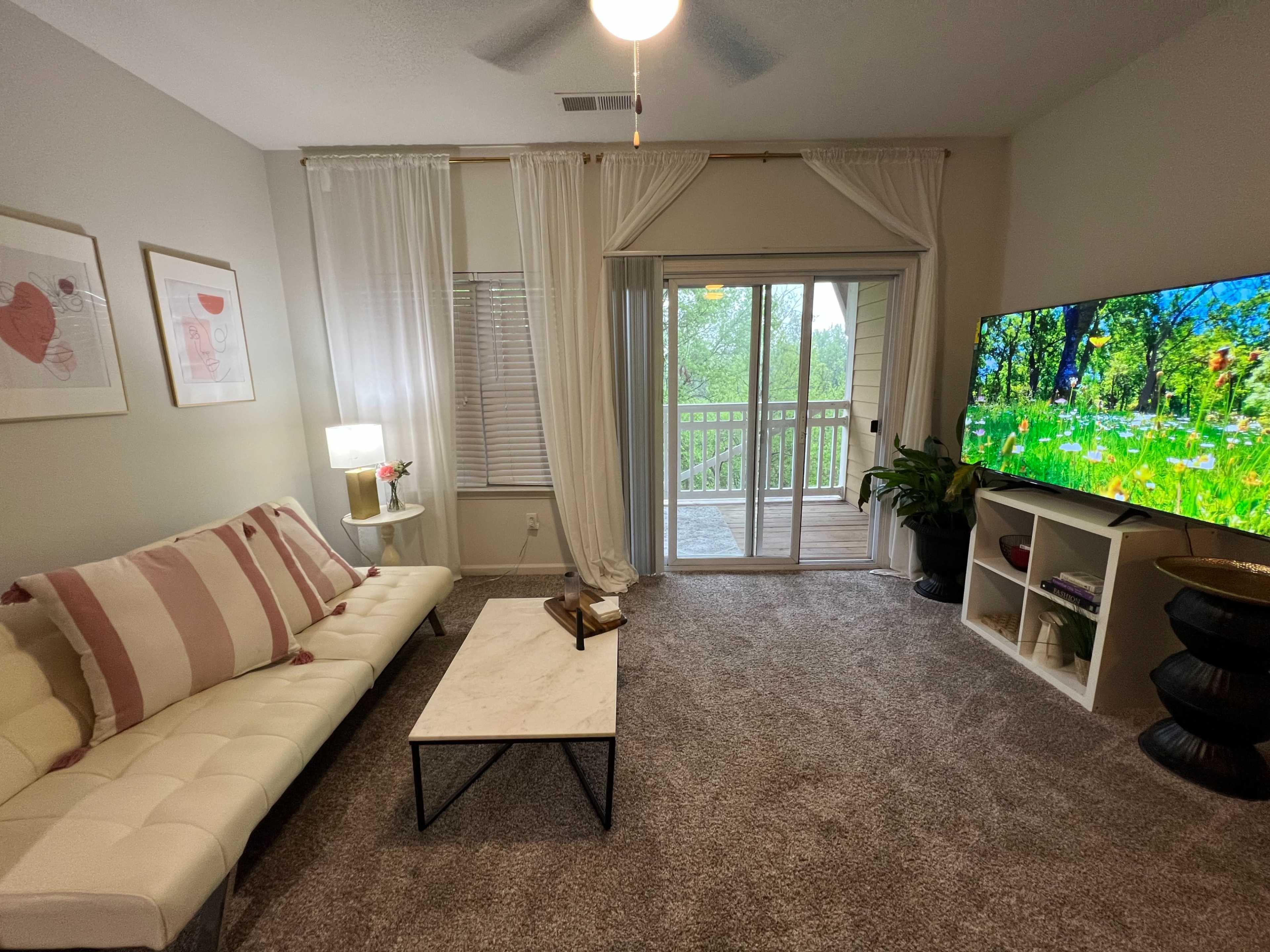 A living room with a white sectional sofa, a marble-topped coffee table, a television on a stand, and a balcony door that opens to a view of greenery.