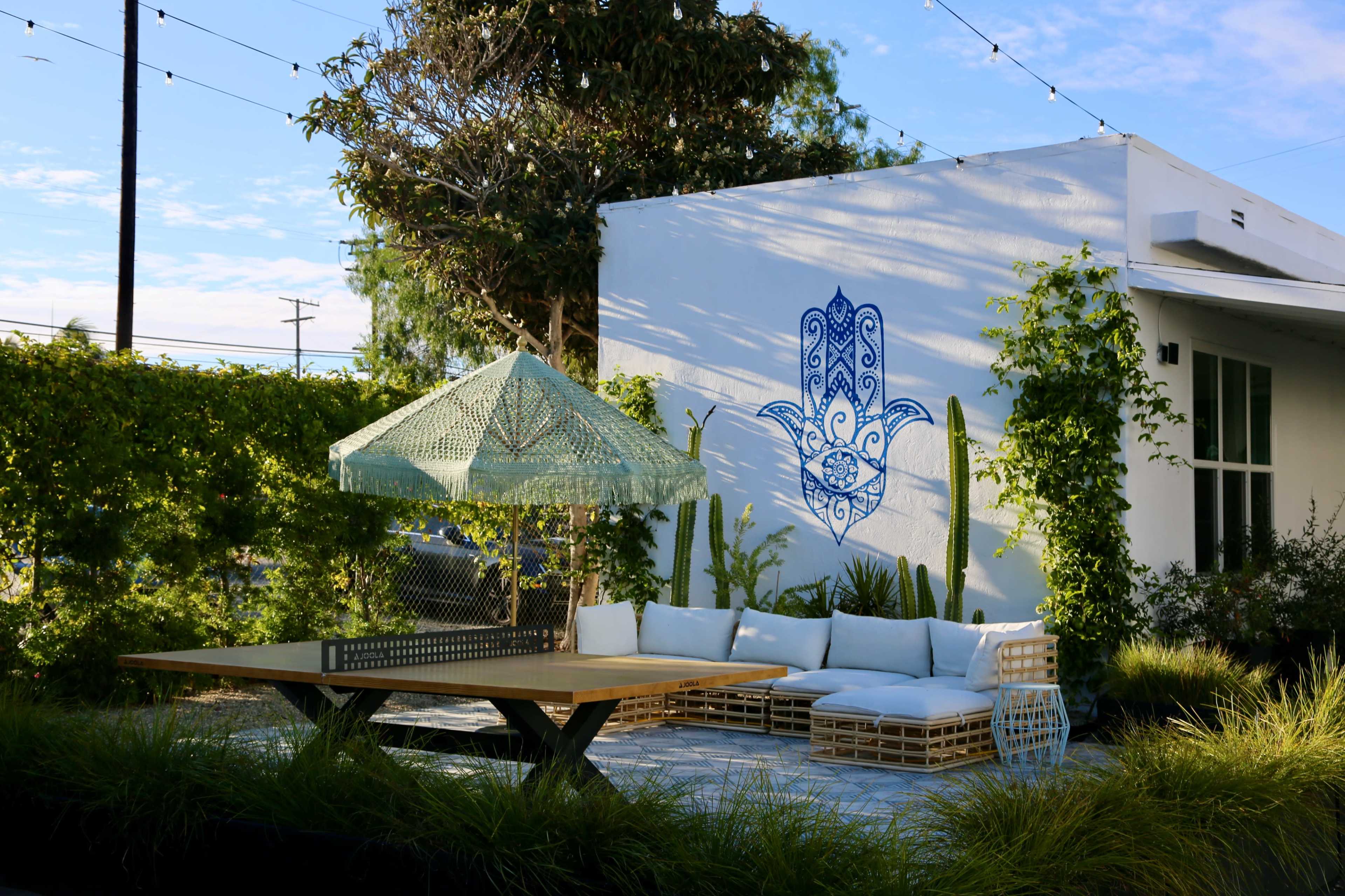 The image features a modern outdoor seating area with a wooden table, white cushions, and a patterned canopy, surrounded by greenery and a decorated wall.