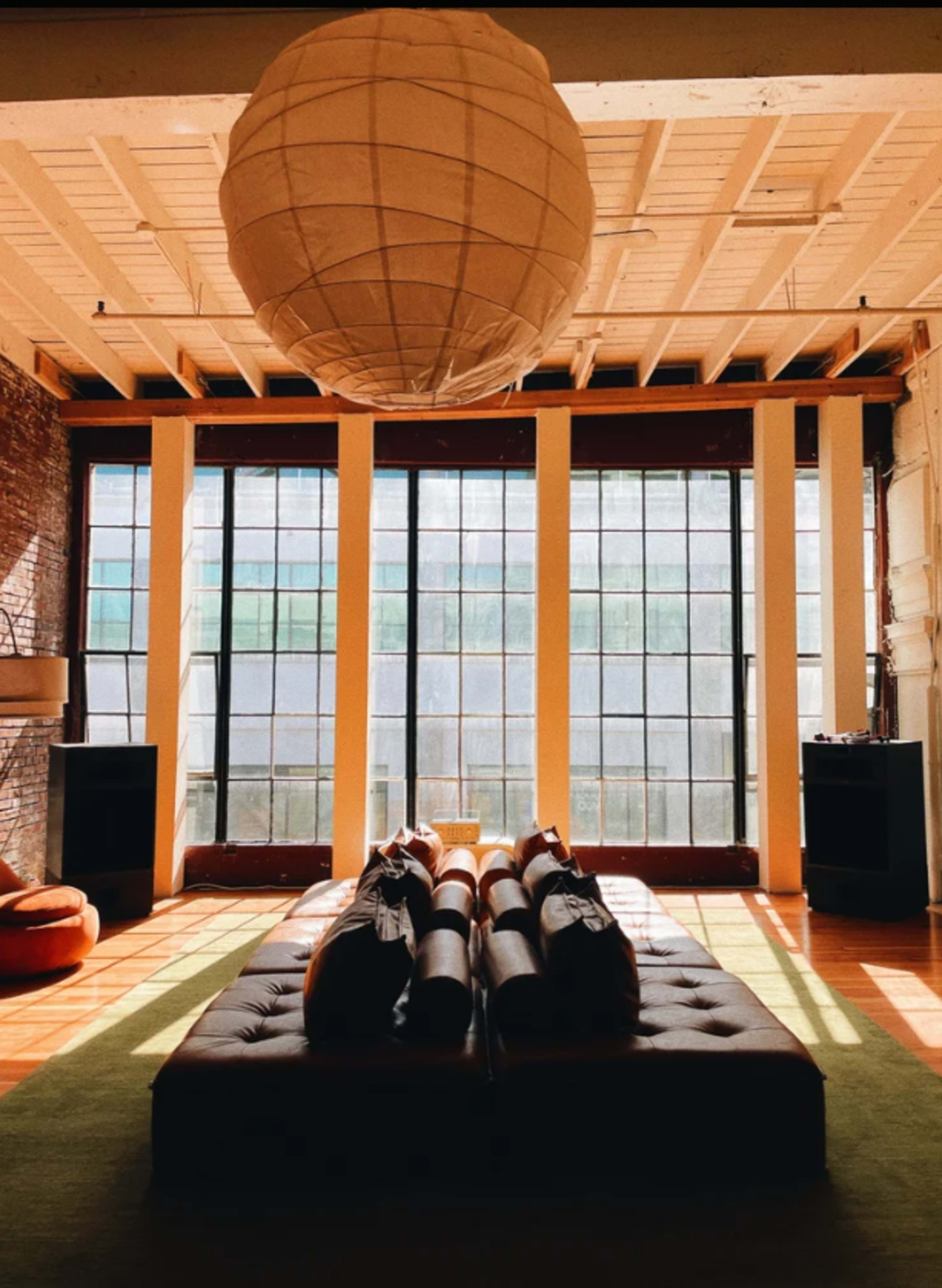 The image shows a spacious room with large windows, wooden flooring, a central sofa arrangement, and a hanging light fixture.