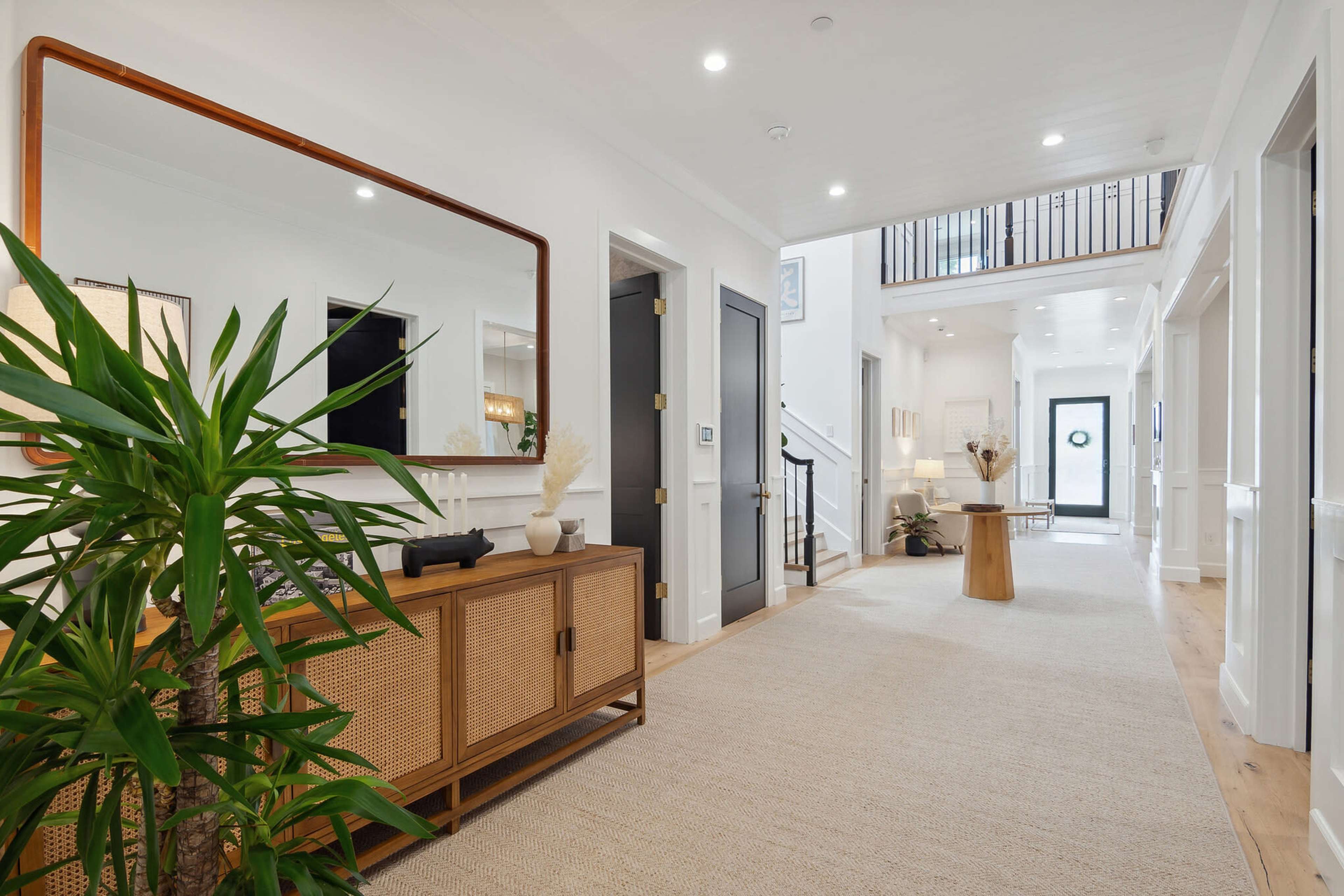 A bright hallway features a large mirror above a console table and leads to a staircase and a door that provides access to the outdoors.