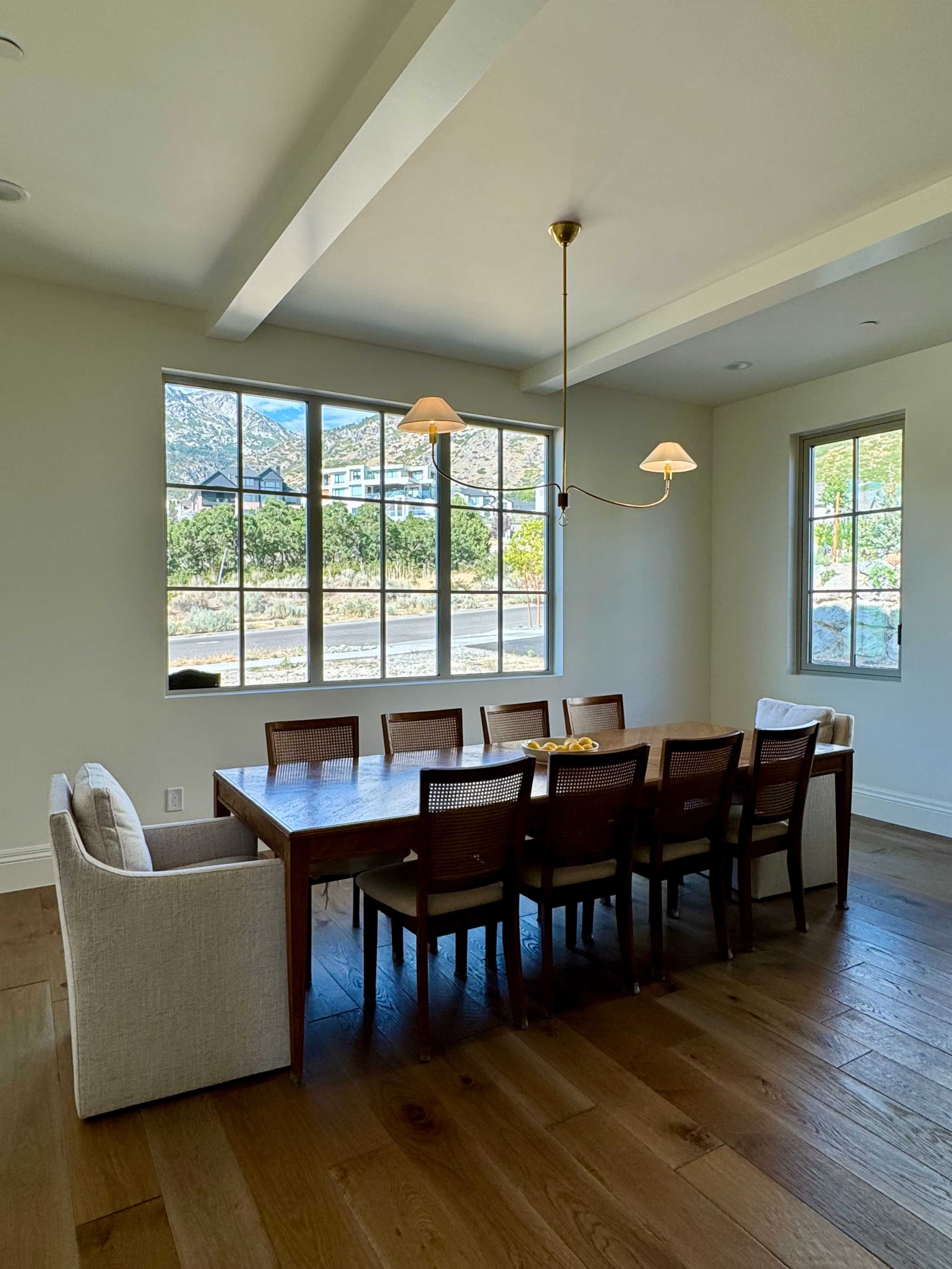The image shows a modern dining room featuring a long wooden table surrounded by eight chairs, with large windows providing a view of the outdoors.