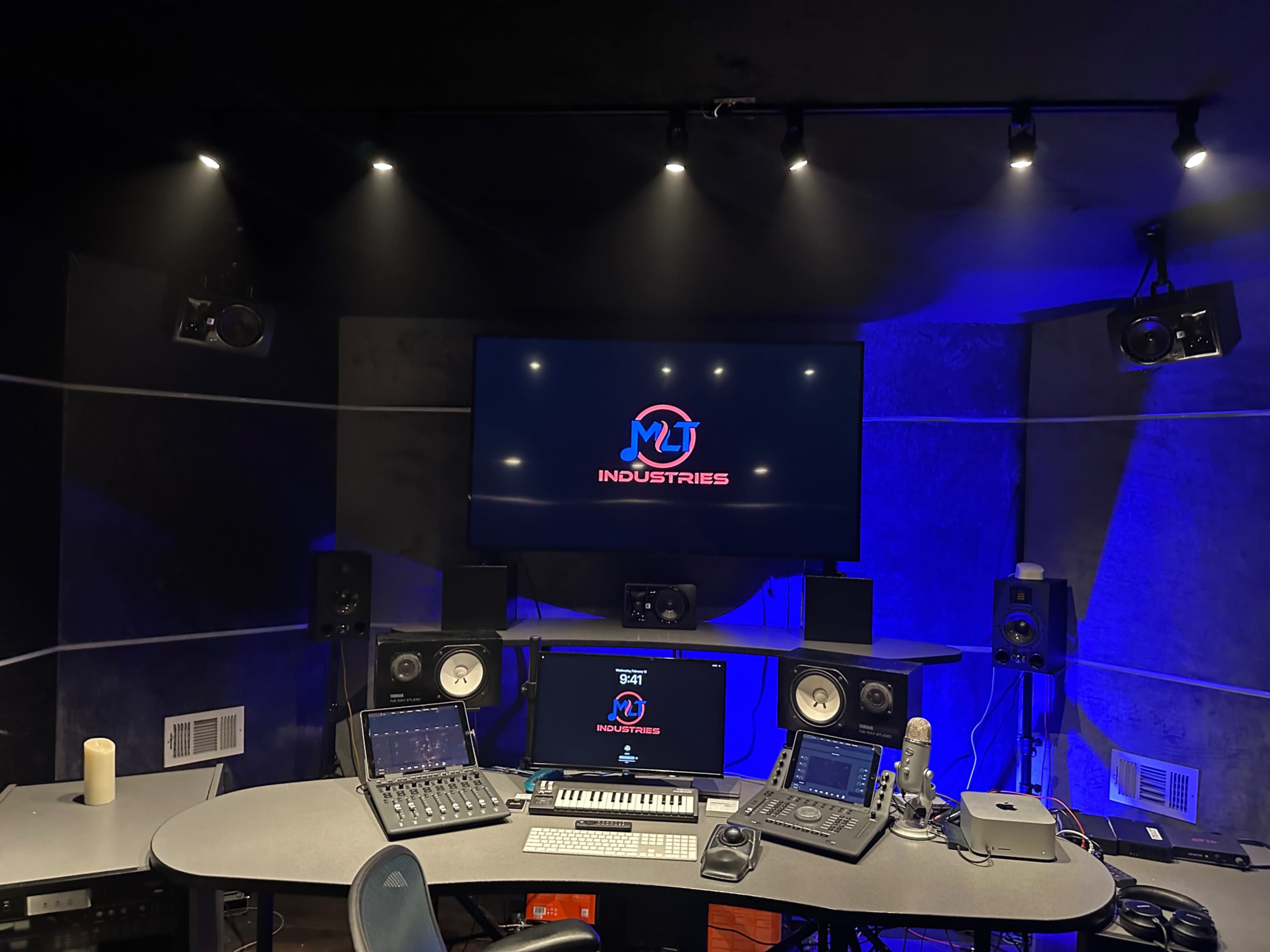 A music studio features a central mixing desk, two large speakers, and a wall-mounted screen displaying the logo "MUT INDUSTRIES."