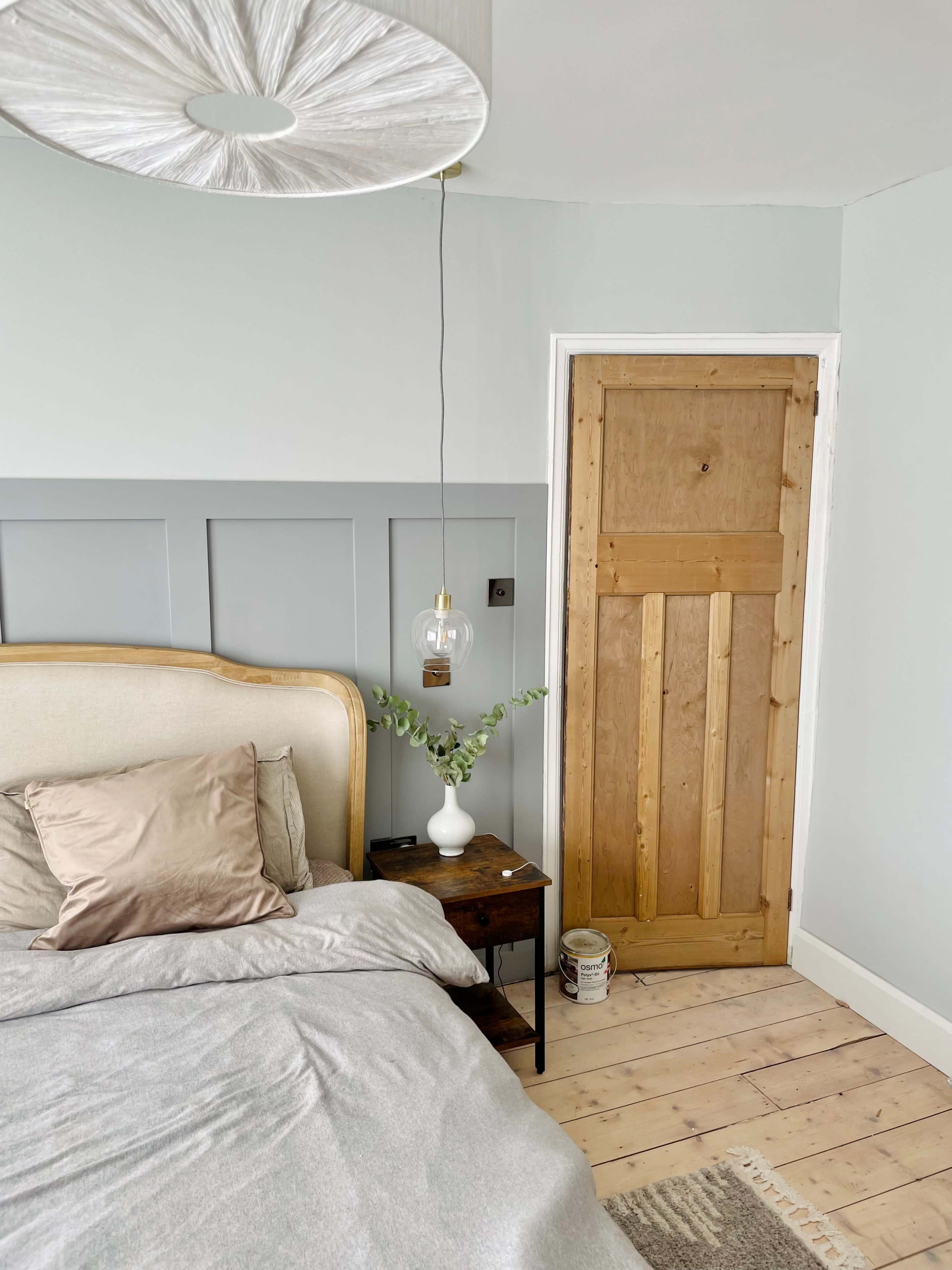 A softly lit bedroom featuring a bed with a beige headboard, a small side table, and a wooden door in a muted color scheme.