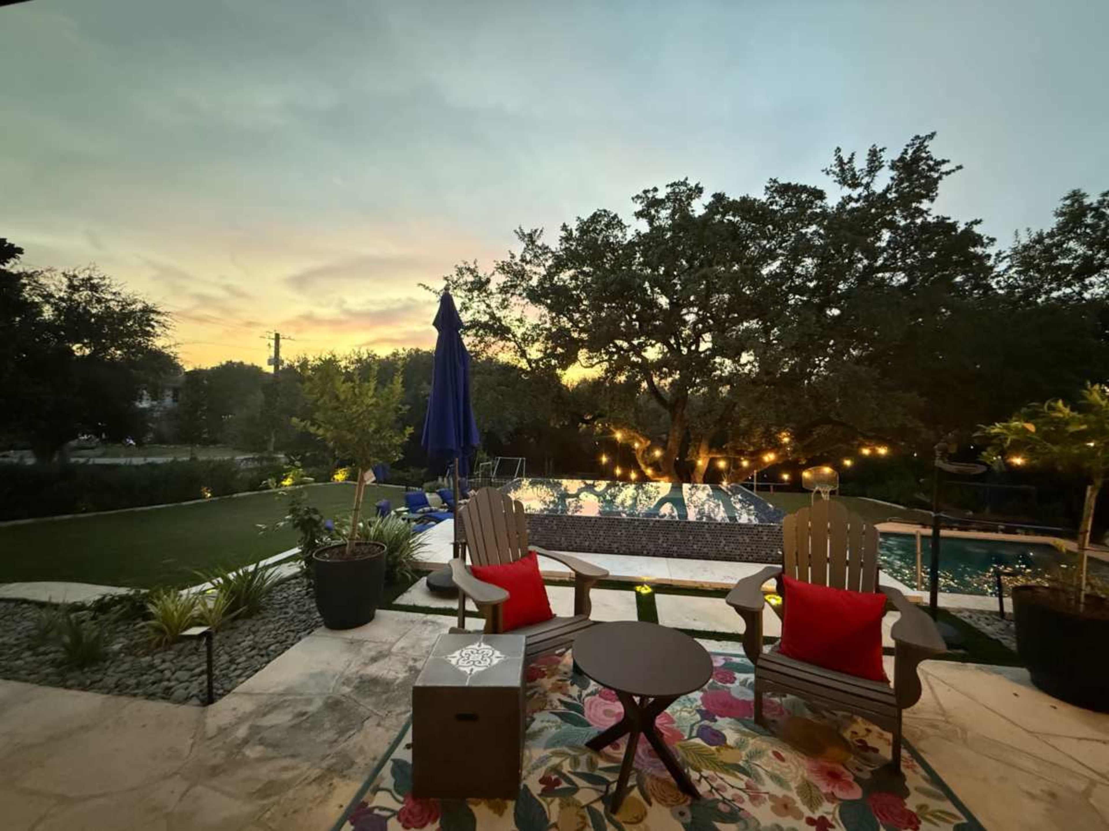The image shows a backyard with two wooden chairs, a small table, a swimming pool, and a sunset in the background, surrounded by greenery and string lights.