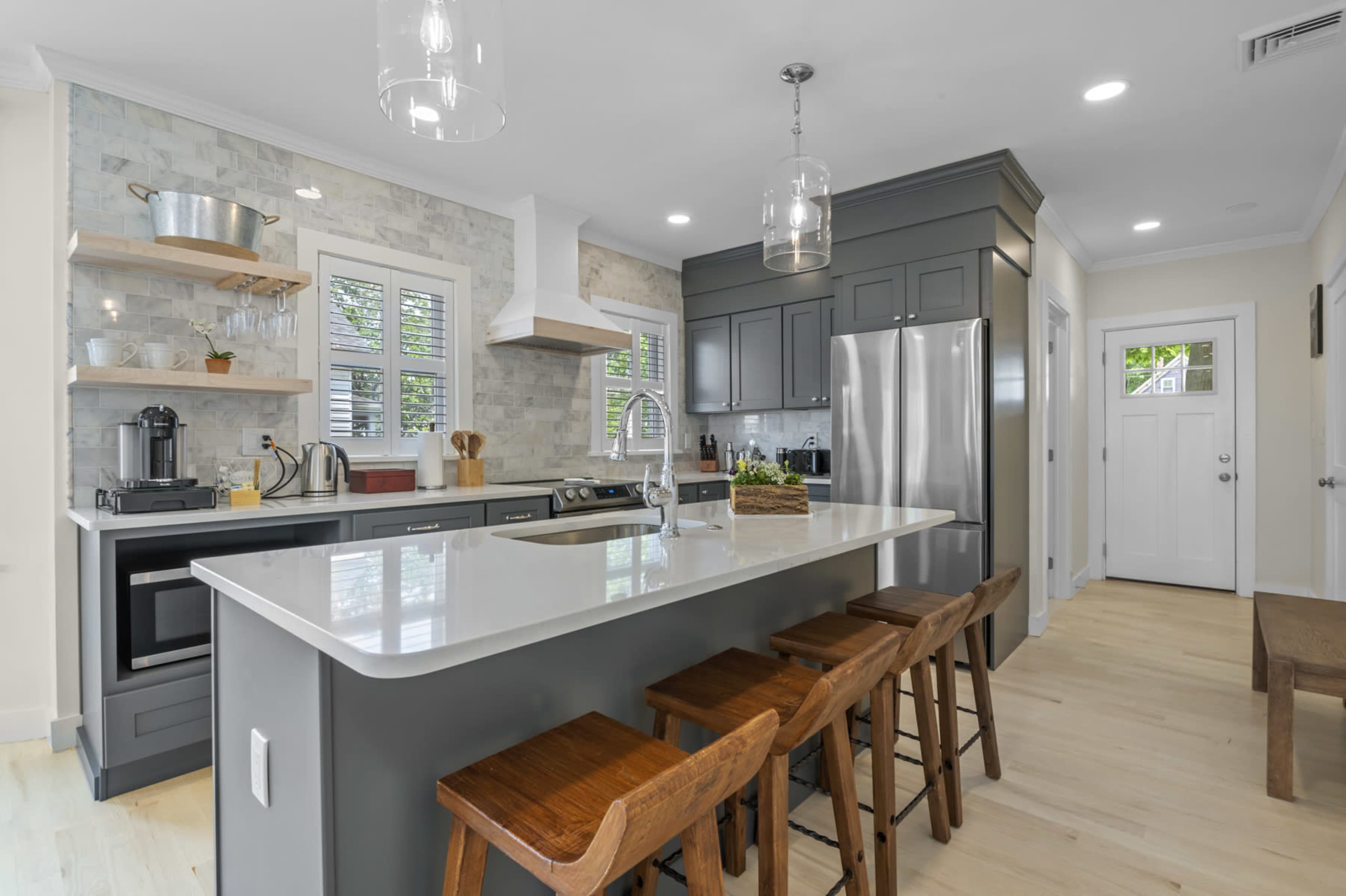 The image shows a modern kitchen featuring gray cabinets, a central island with bar stools, and stainless steel appliances.