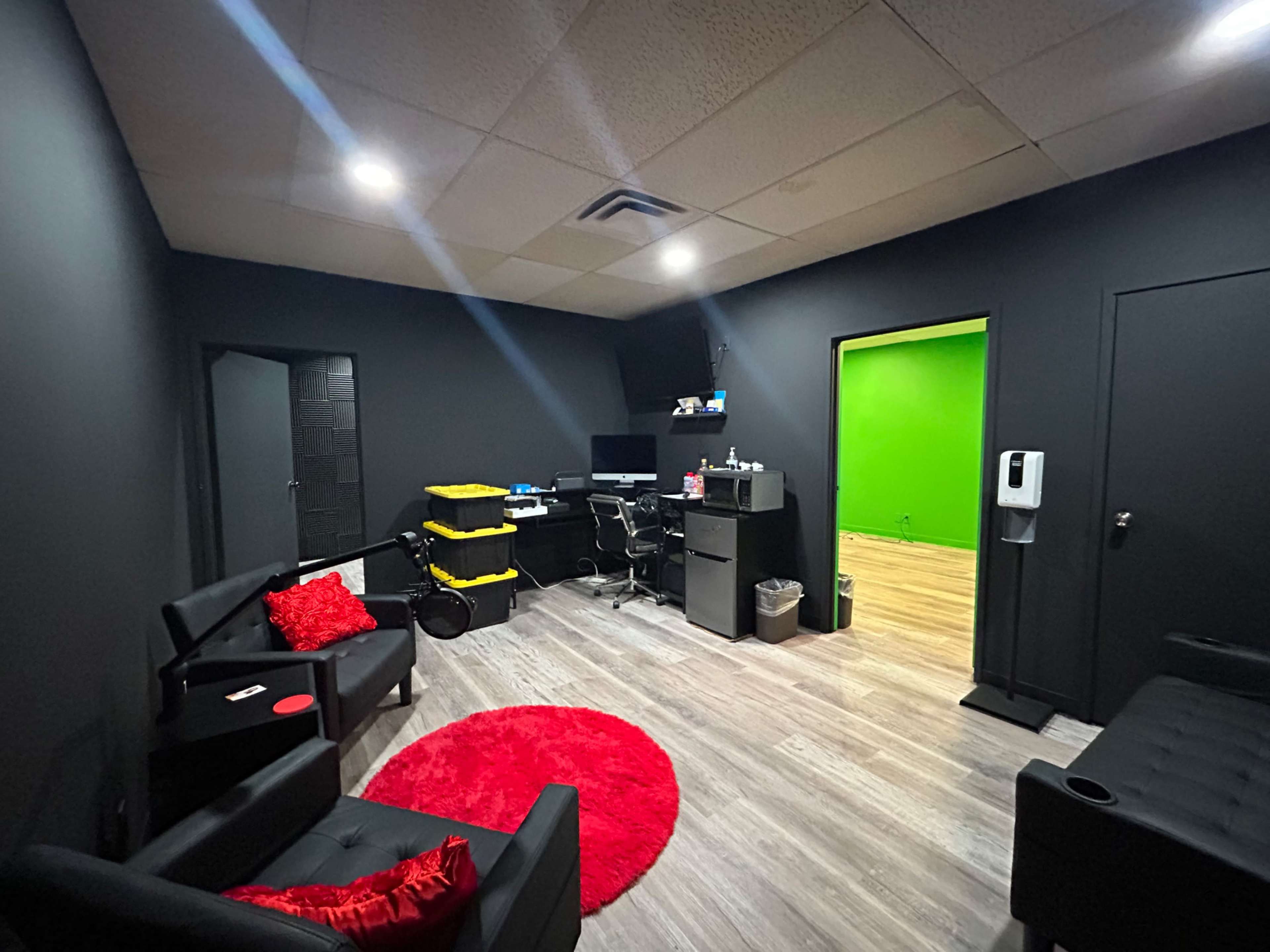 The image shows a dark-colored room with modern furnishings, including black sofas, a small round red rug, and a workspace with a desk and computer, alongside a bright green wall.