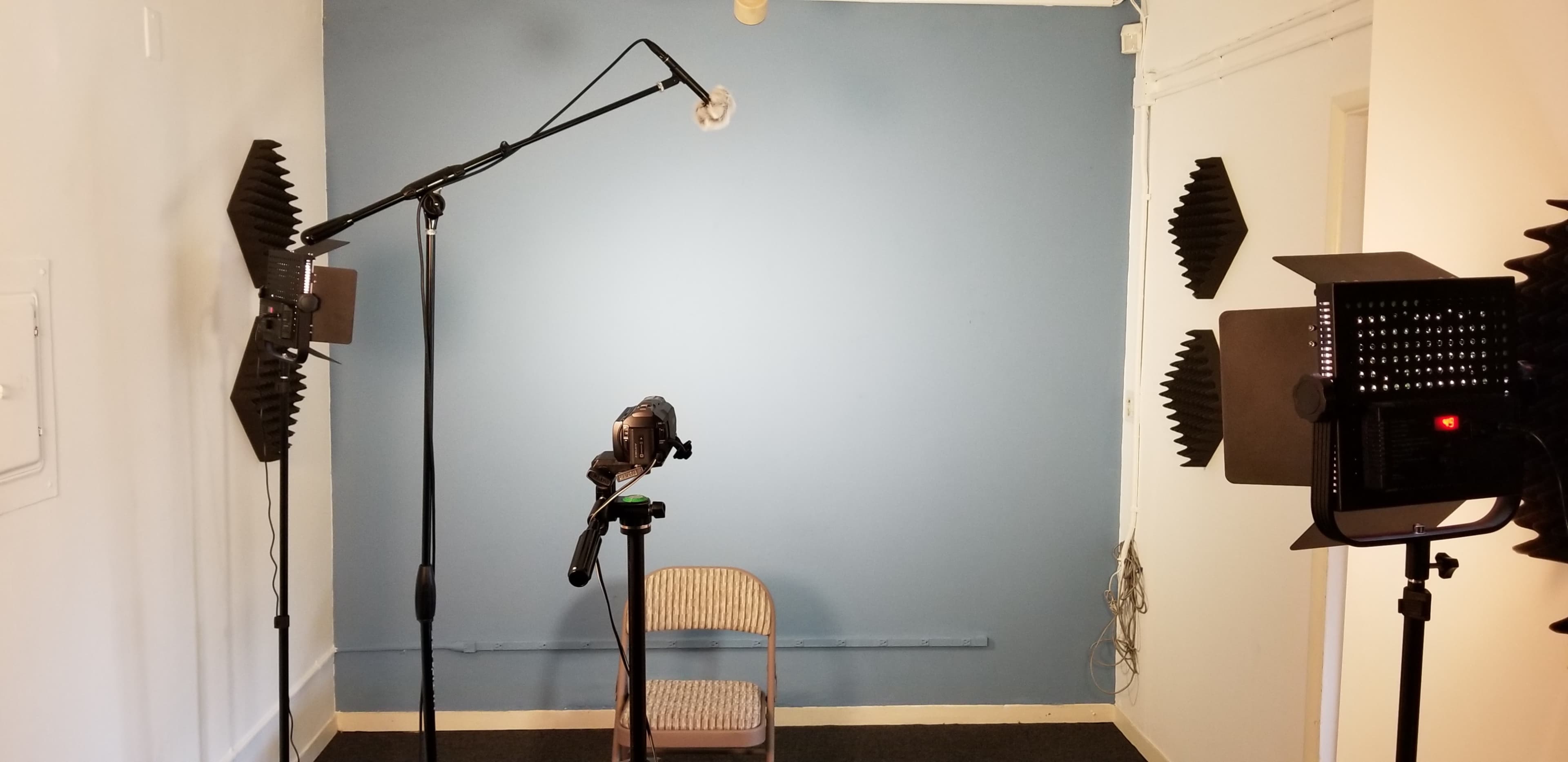 A recording setup in a soundproofed room, featuring a camera on a tripod, a microphone on a stand, and two softbox lights aimed at a blue wall.
