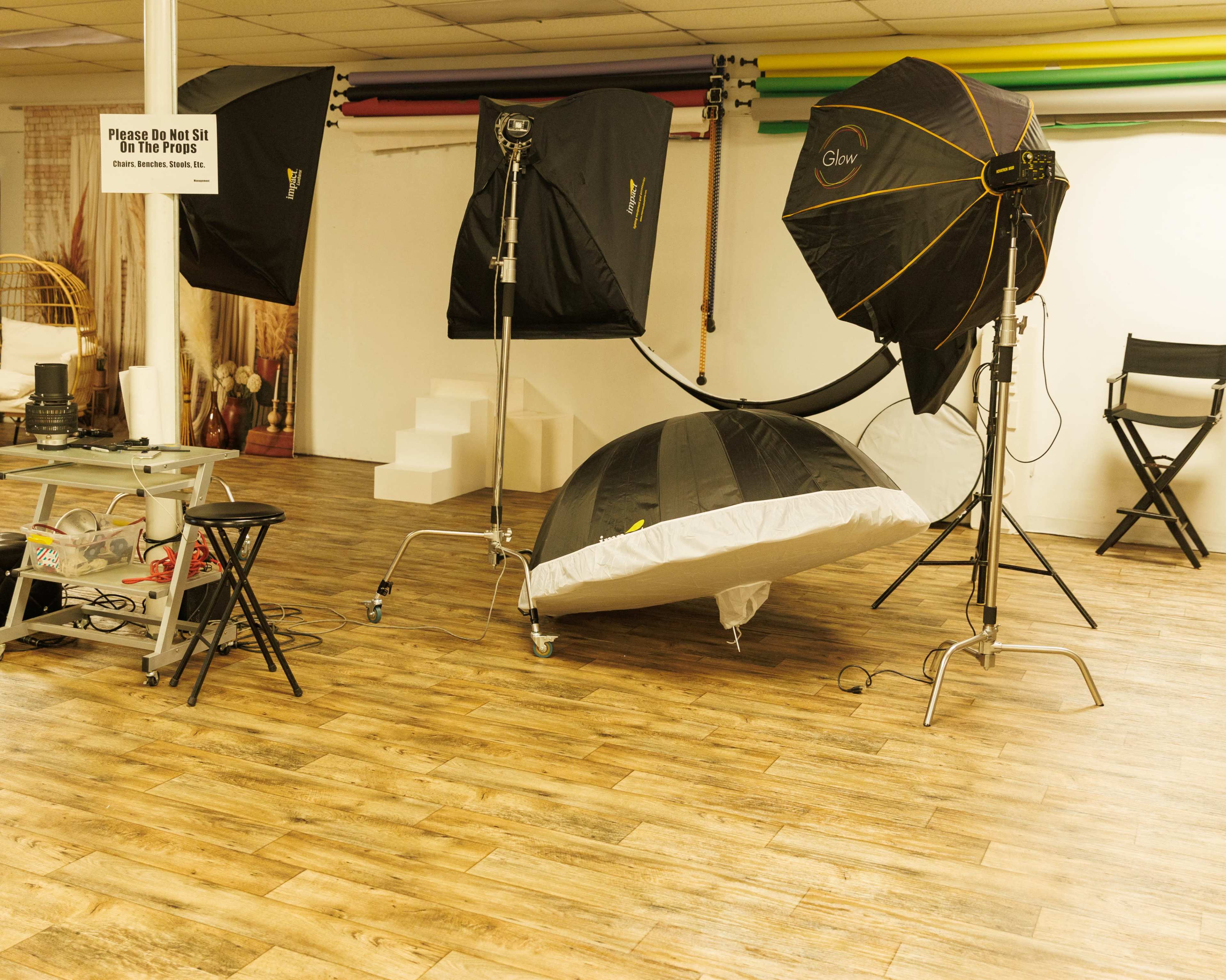 The image shows a photography studio setup with multiple softbox lights, a large reflective umbrella, and a low-profile bed on a wood-finish floor.
