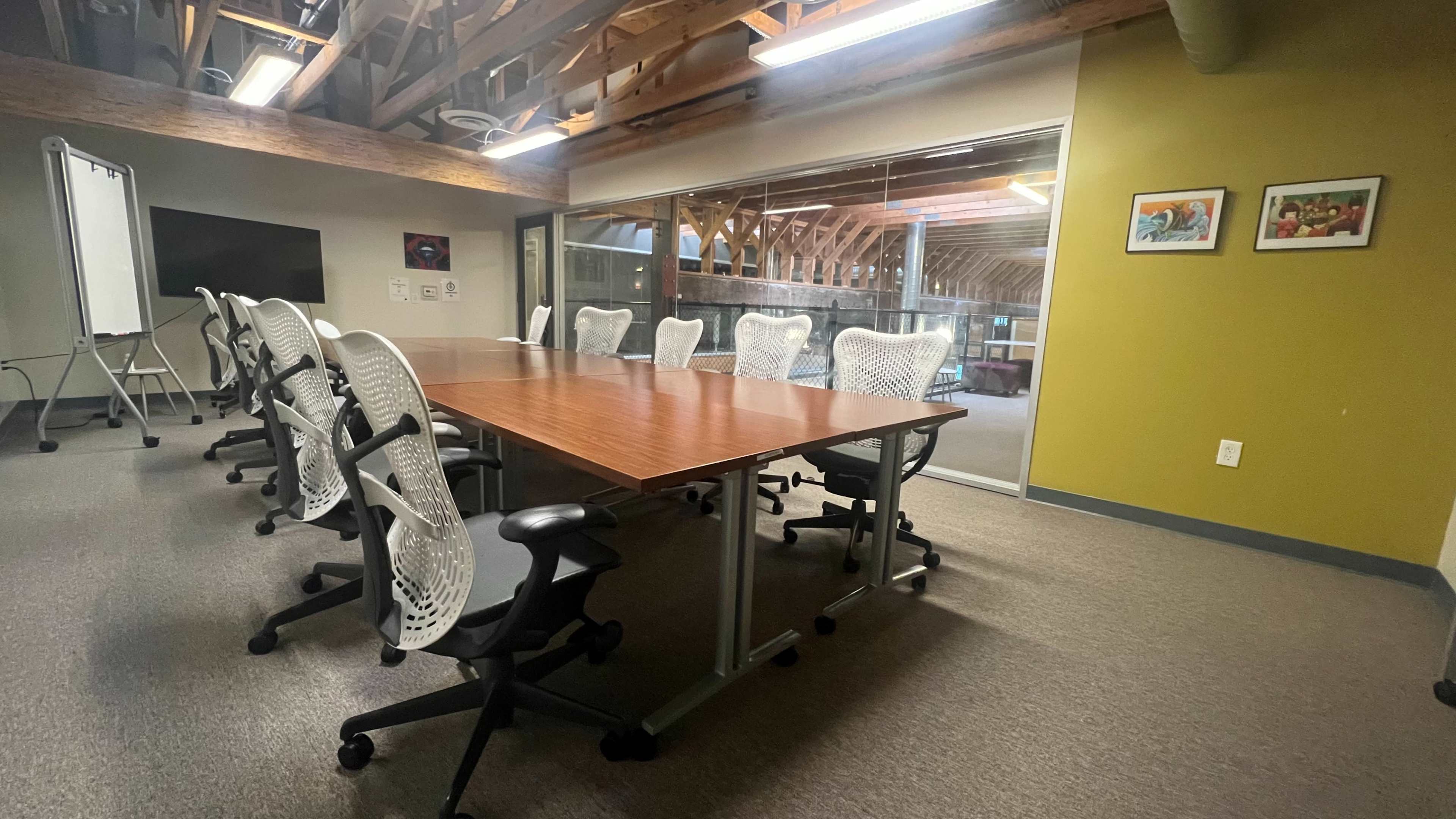 A conference room is set up with a large wooden table surrounded by office chairs, with a whiteboard and wall-mounted screen visible in the background.
