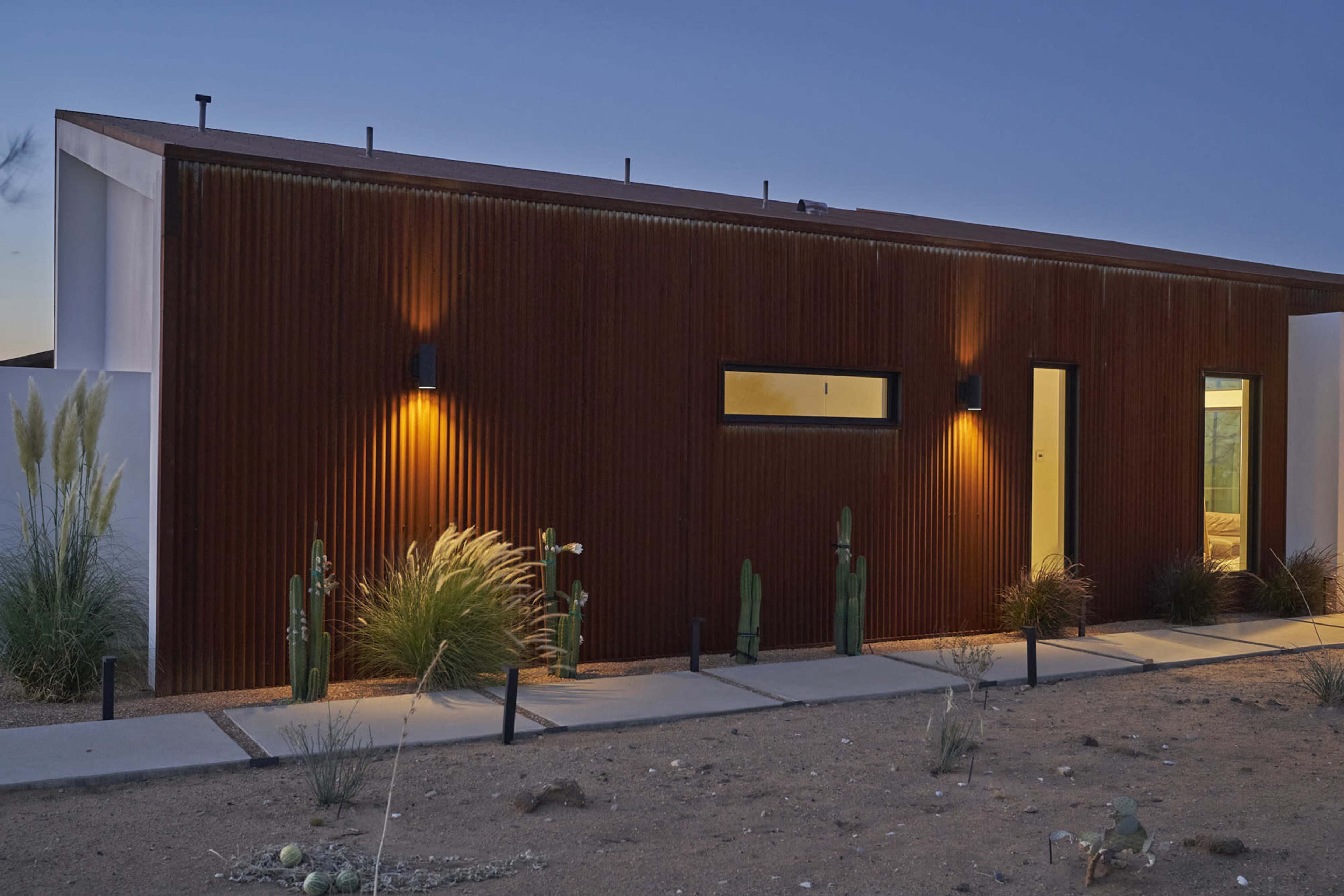 The image shows a modern house with a corrugated metal exterior, side lighting, and desert landscaping at twilight.
