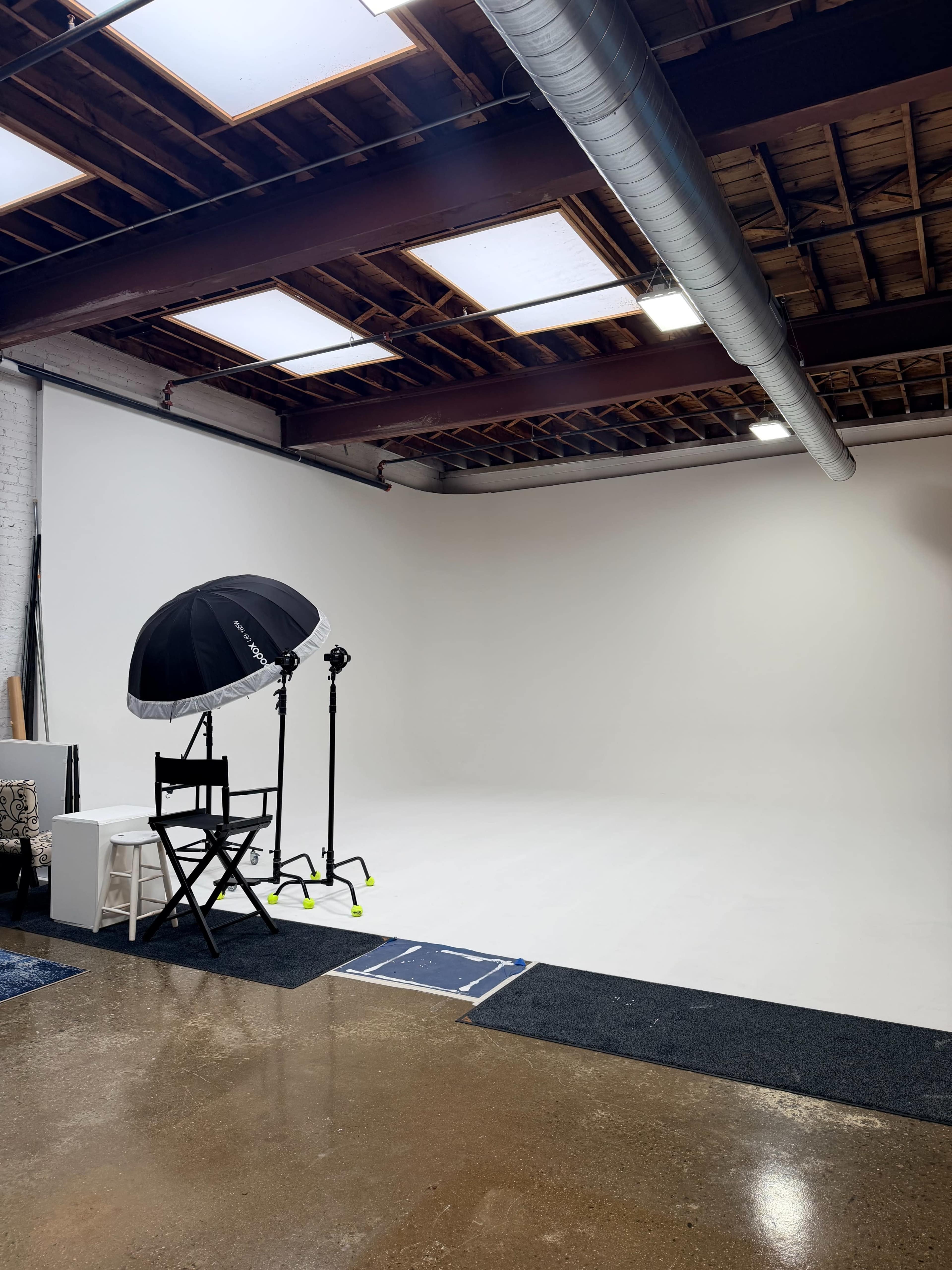 The image shows a photo studio with a white backdrop, a director's chair, and an umbrella light setup.