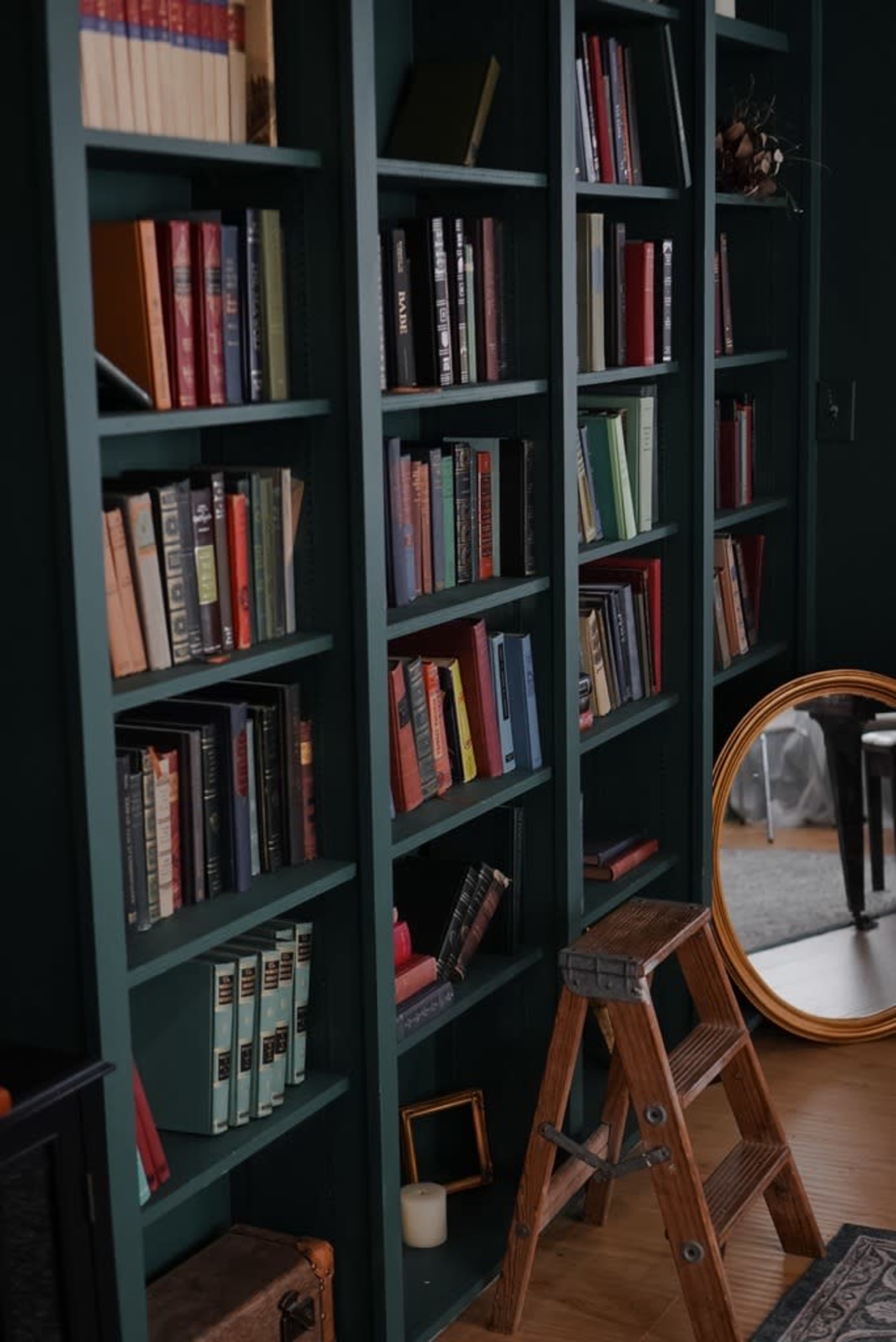The image shows a tall bookshelf filled with various books, accompanied by a wooden step ladder and a round mirror.