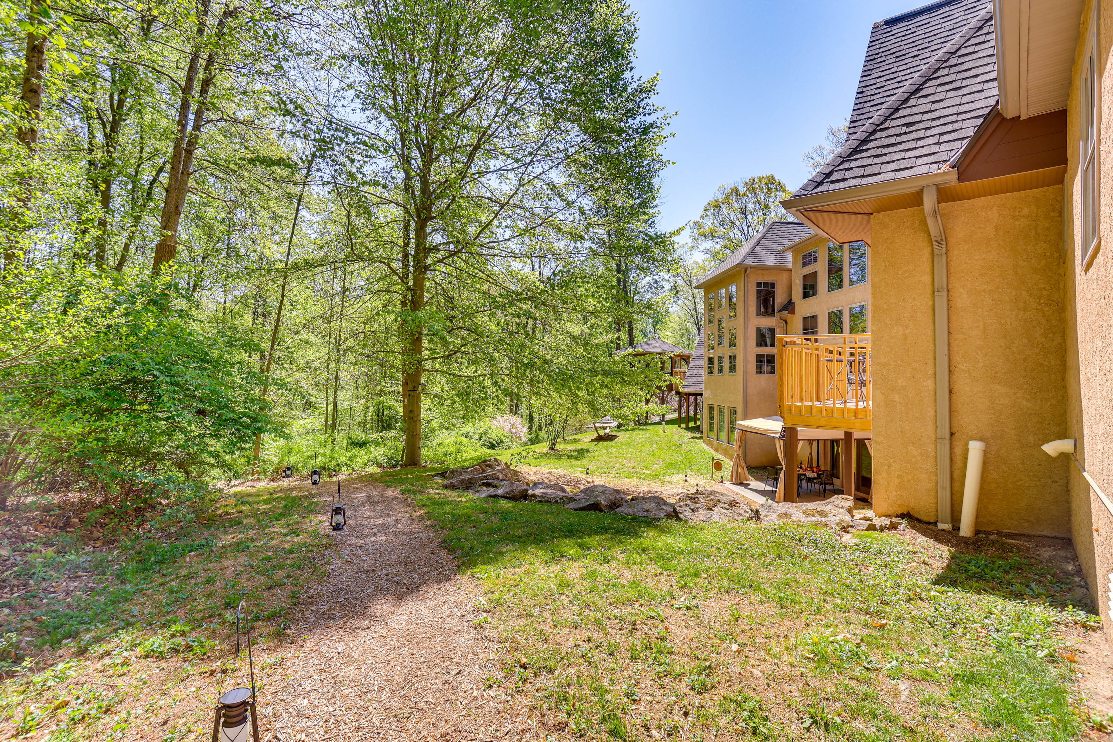 A pathway lined with natural grass and rocks leads past a house surrounded by trees and greenery.