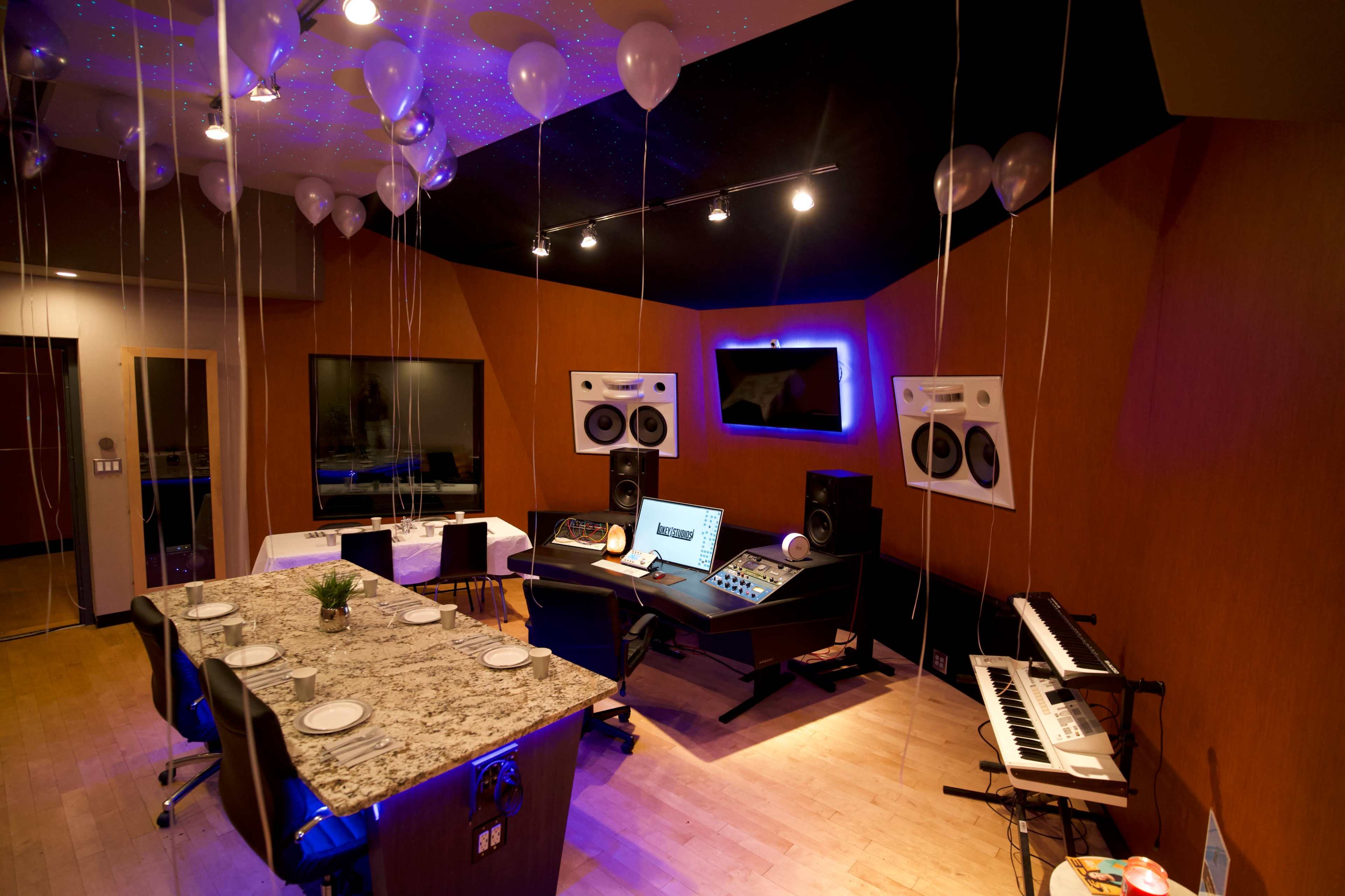 A modern music studio featuring a central mixing console, audio speakers, a keyboard, and a dining table surrounded by balloons and softly illuminated ceiling lights.