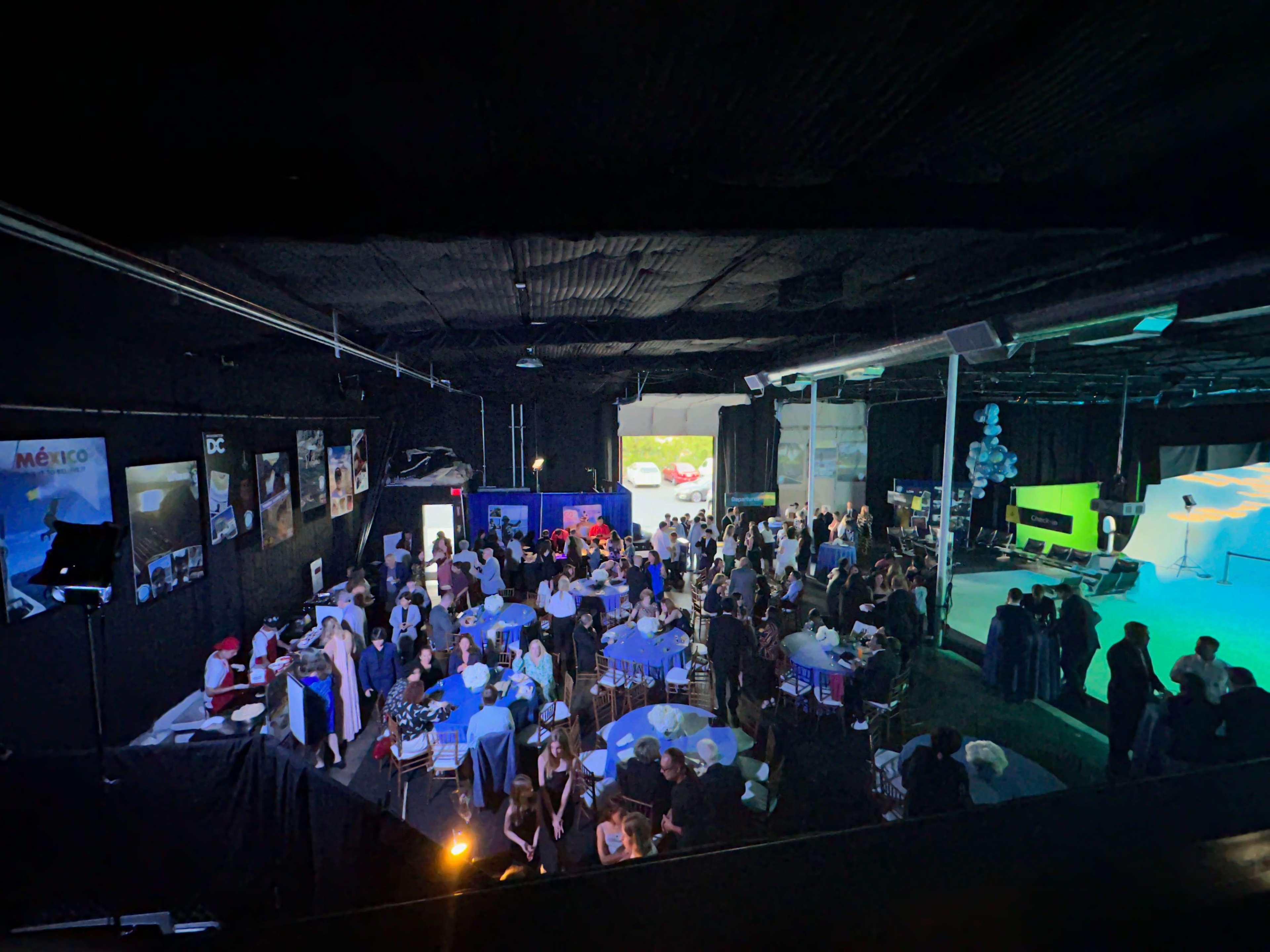 A large gathering of people is taking place inside a dark, spacious venue, featuring tables with blue tablecloths and decorations, while a stage and screens are set up in the background.